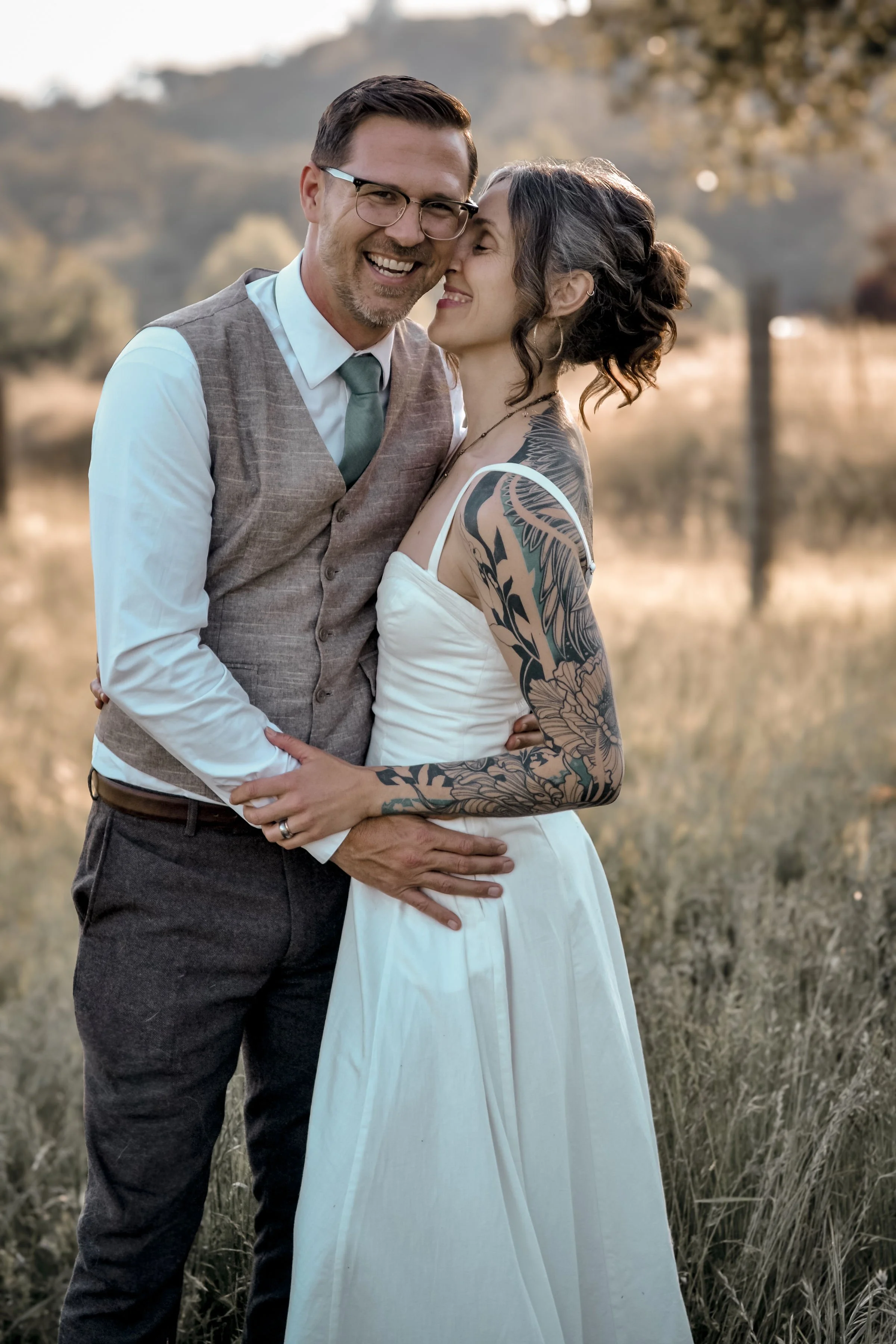 A smiling couple embracing outdoors in a field with a blurred natural landscape in the background, the man wearing glasses, a white shirt, and vest, and the woman with tattoos on her arm and wearing a white dress.