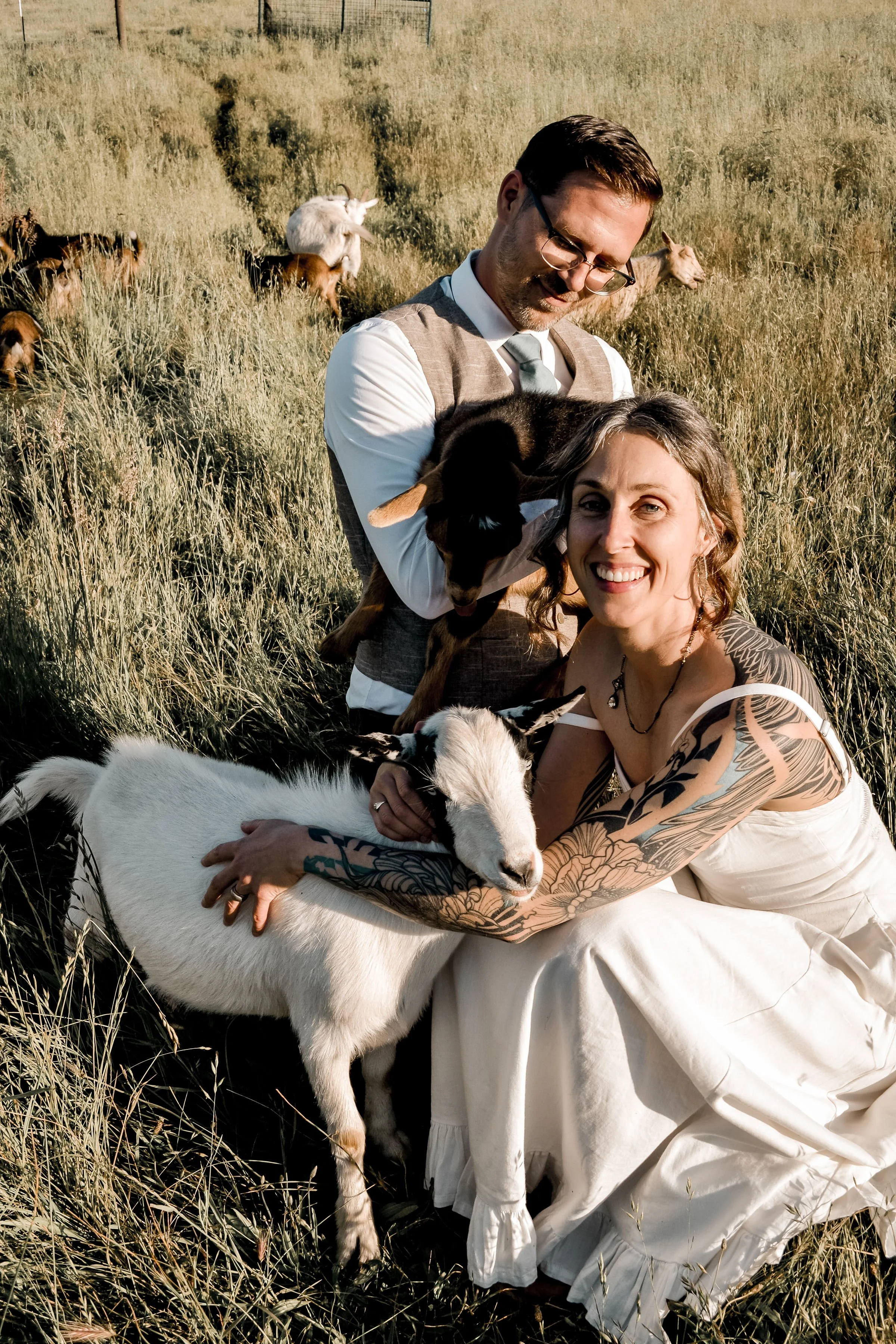 A man and a woman with tattoos sitting on the grass in a field, holding and hugging goats, with more goats in the background.