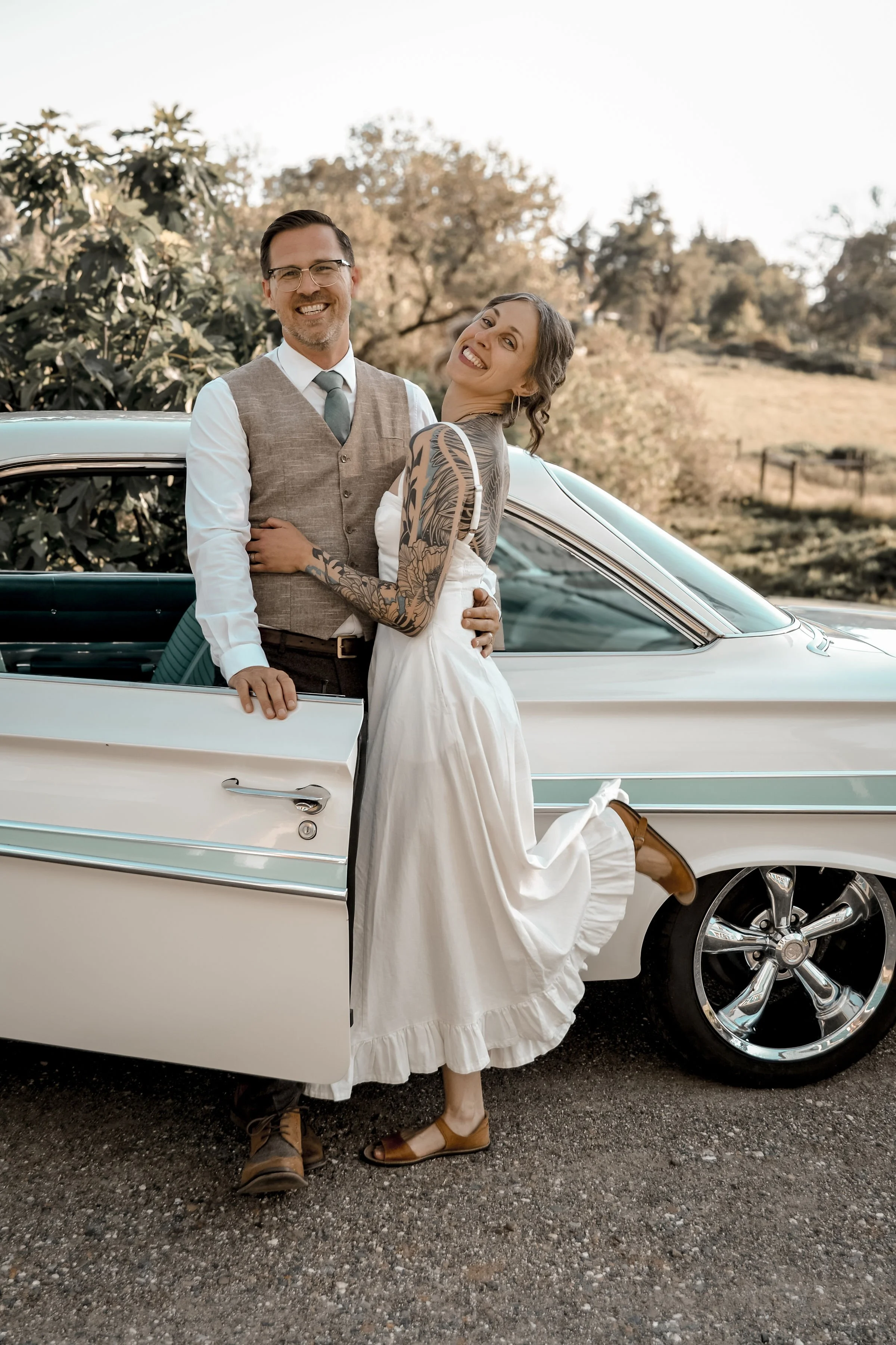 A smiling man in a vest and tie and a woman in a white dress with tattoos on her arms and legs, leaning against a vintage white car parked outdoors on a gravel road, with a scenic background of trees and hills.