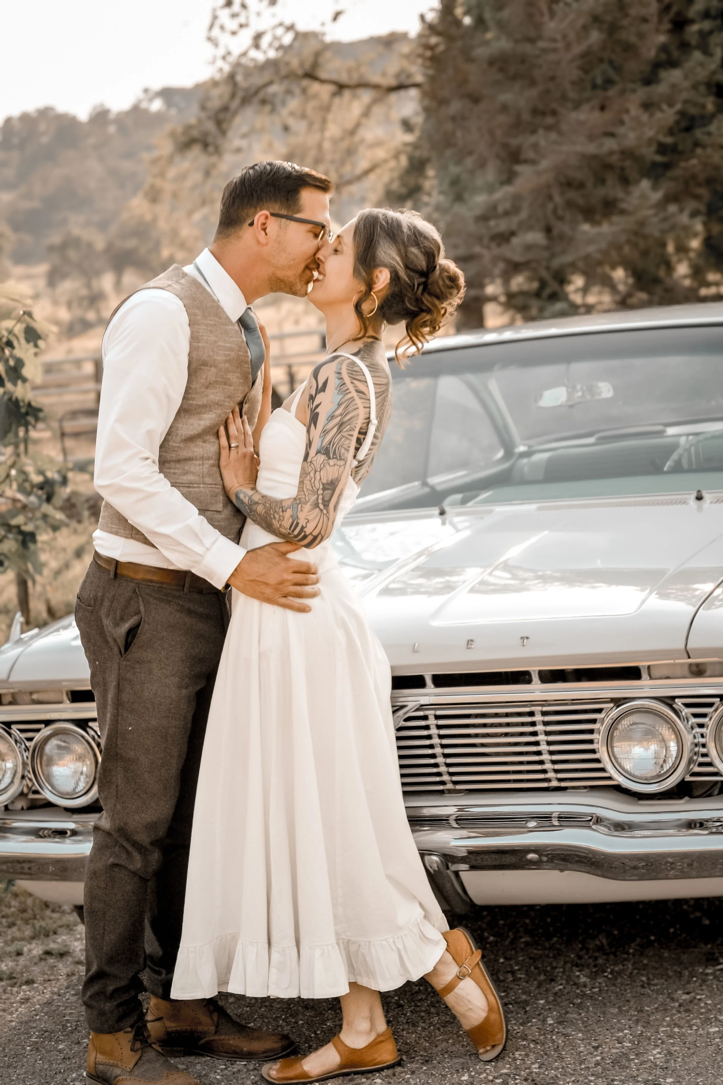 A couple is kissing in front of a vintage Cadillac car outdoors on a sunny day. The man is wearing glasses, a white shirt, a gray vest, and brown pants with boots. The woman has tattoos on her arms, her hair in an updo, and is wearing a white dress w