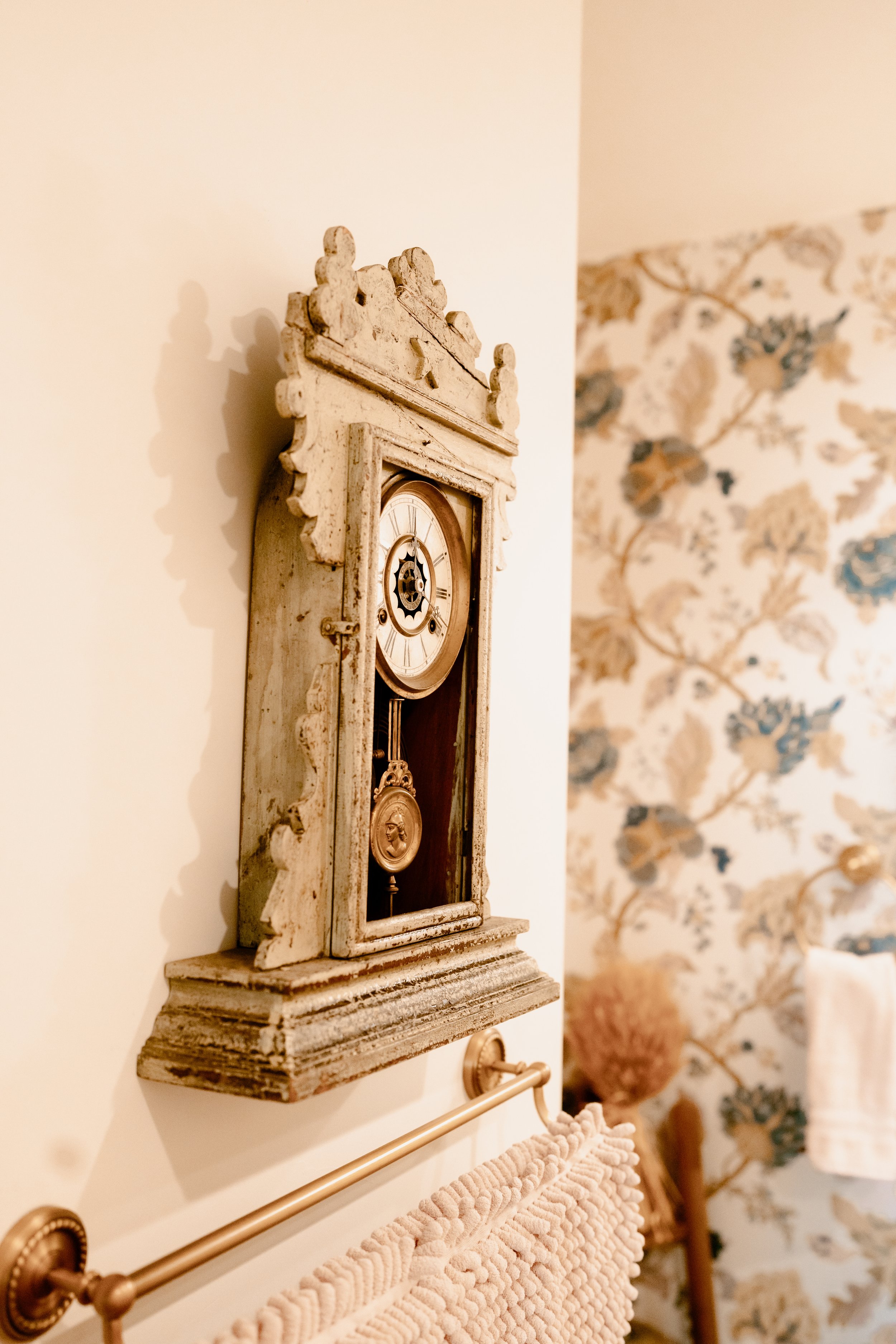 Close-up of a vintage wall clock with a distressed white finish, mounted on a wallpapered wall with a floral pattern.