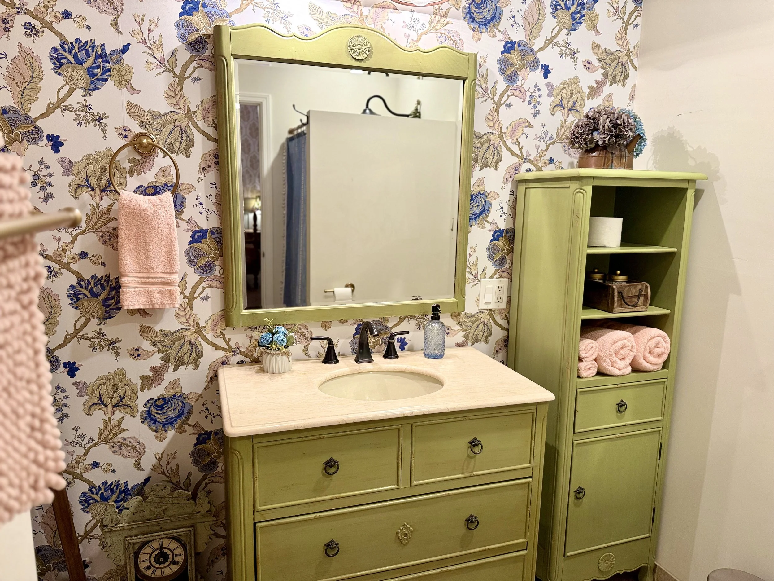 A bathroom with floral wallpaper, a lime green vanity with a mirror, a towel ring with a pink hand towel, a green cabinet with pink towels, a clock, and various small decor items.