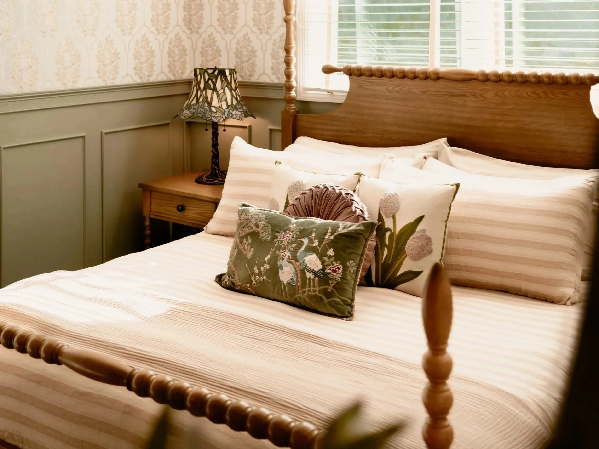 A bedroom scene featuring a wooden bed with pillows, a Tiffany-style stained glass lamp on a wooden nightstand, and a window with blinds. The wall has patterned wallpaper and wood paneling.