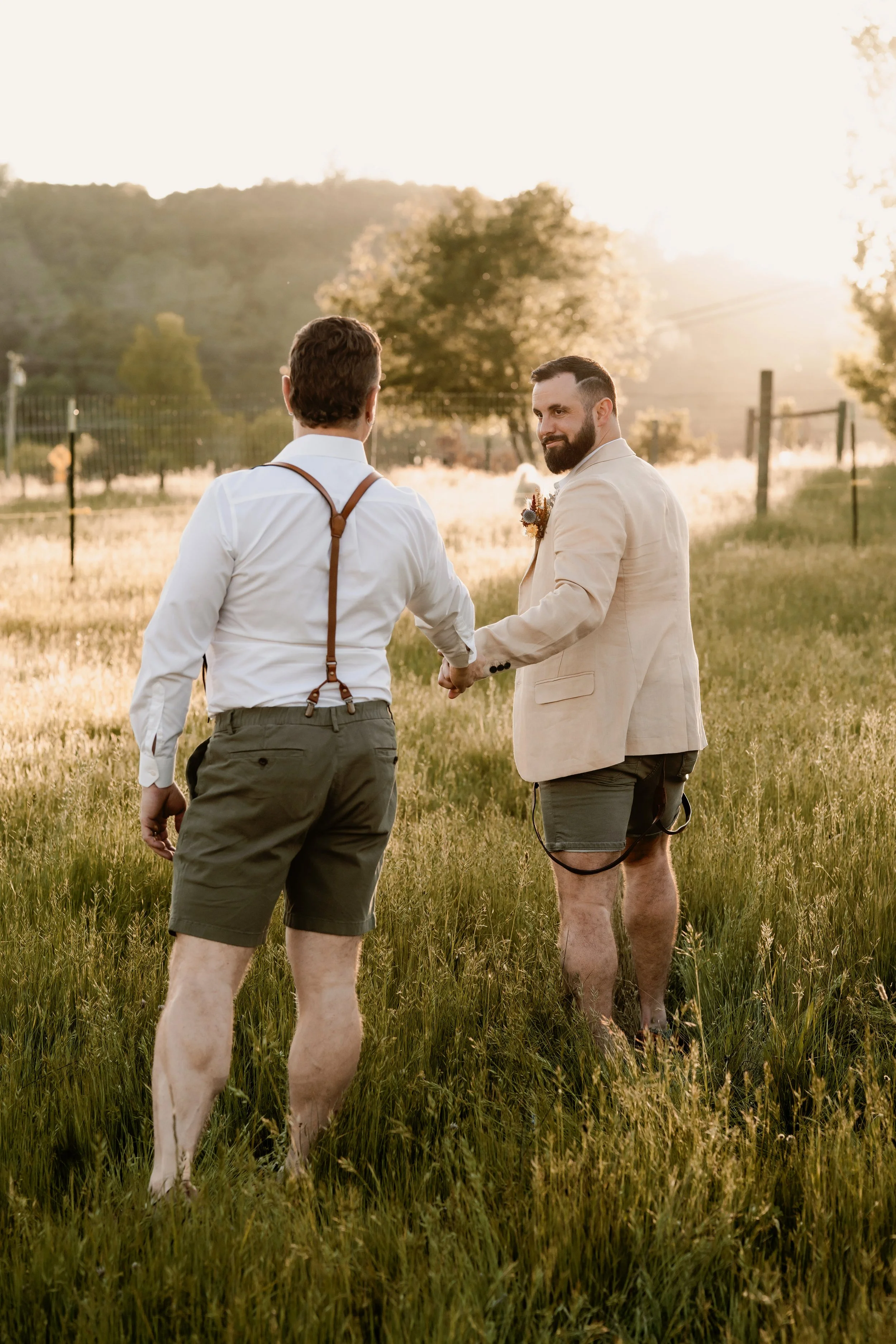 Two men holding hands in a field during sunset, one dressed in a beige blazer and shorts with a beard, and the other in a white shirt with suspenders and khaki shorts.