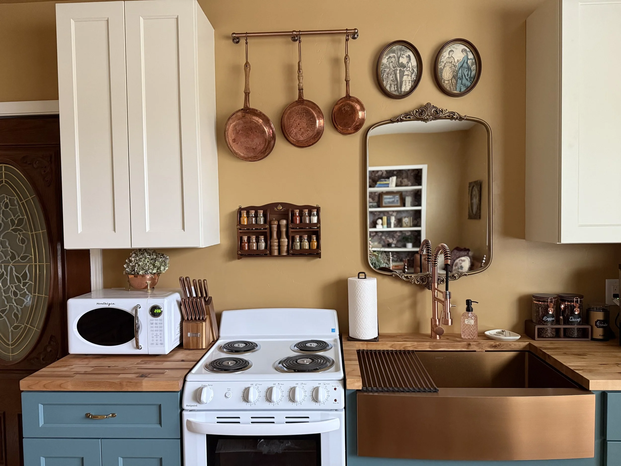 Kitchen countertop with a microwave, a knife block, a stove, and a copper sink. Copper pots hang on the beige wall above, with a mirror and framed pictures behind. Small containers, a paper towel roll, and a soap dispenser are near the sink.