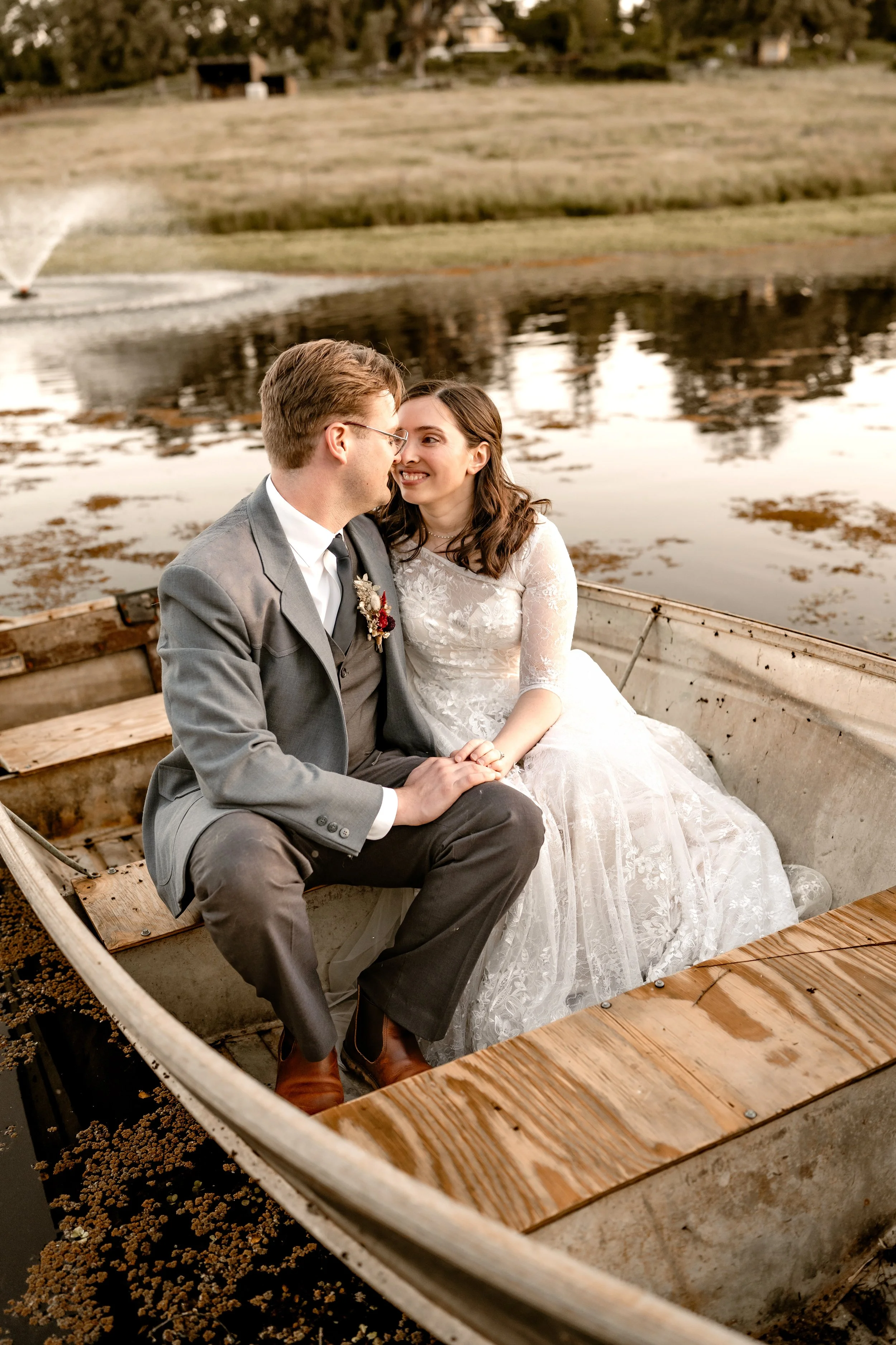 A newlywed couple dressed in wedding attire, sitting in a small boat on a calm lake at sunset, smiling and looking into each other's eyes, with grassy hills and trees in the background.