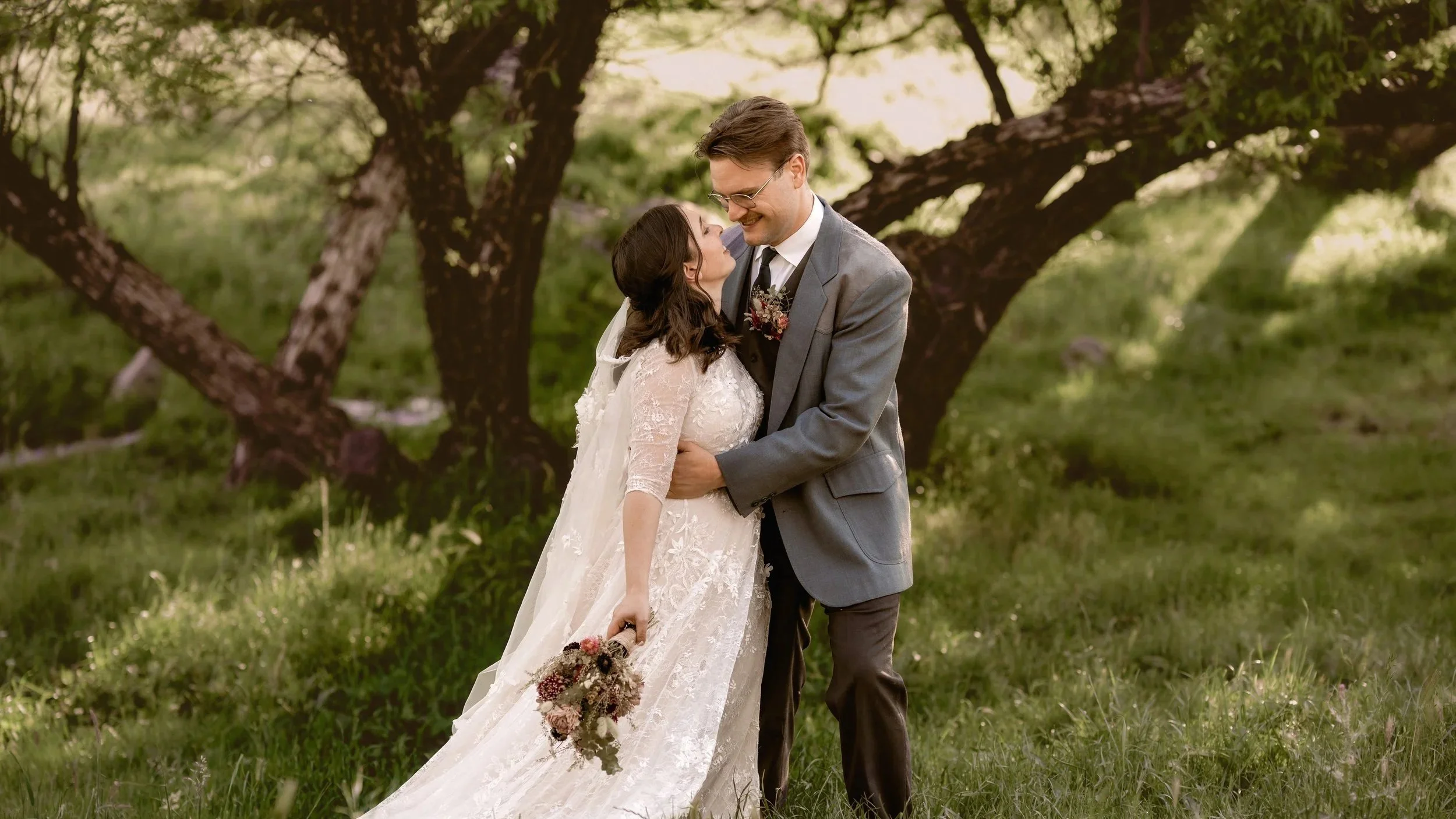 A bride and groom embrace outdoors beneath a tree, smiling at each other. The bride holds a bouquet, and both are dressed in wedding attire.