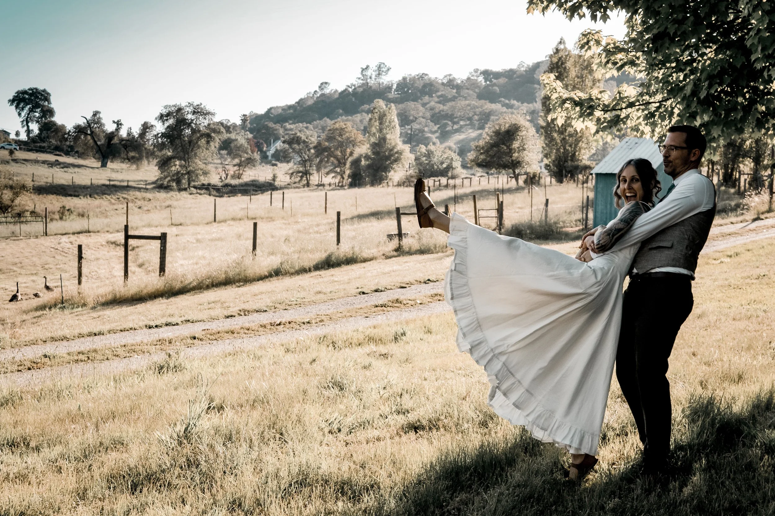 A man is holding a woman in a white dress in a rural outdoor setting with grassy fields, trees, and hills in the background. The woman is smiling and laughing, and the man is wearing glasses and a vest.