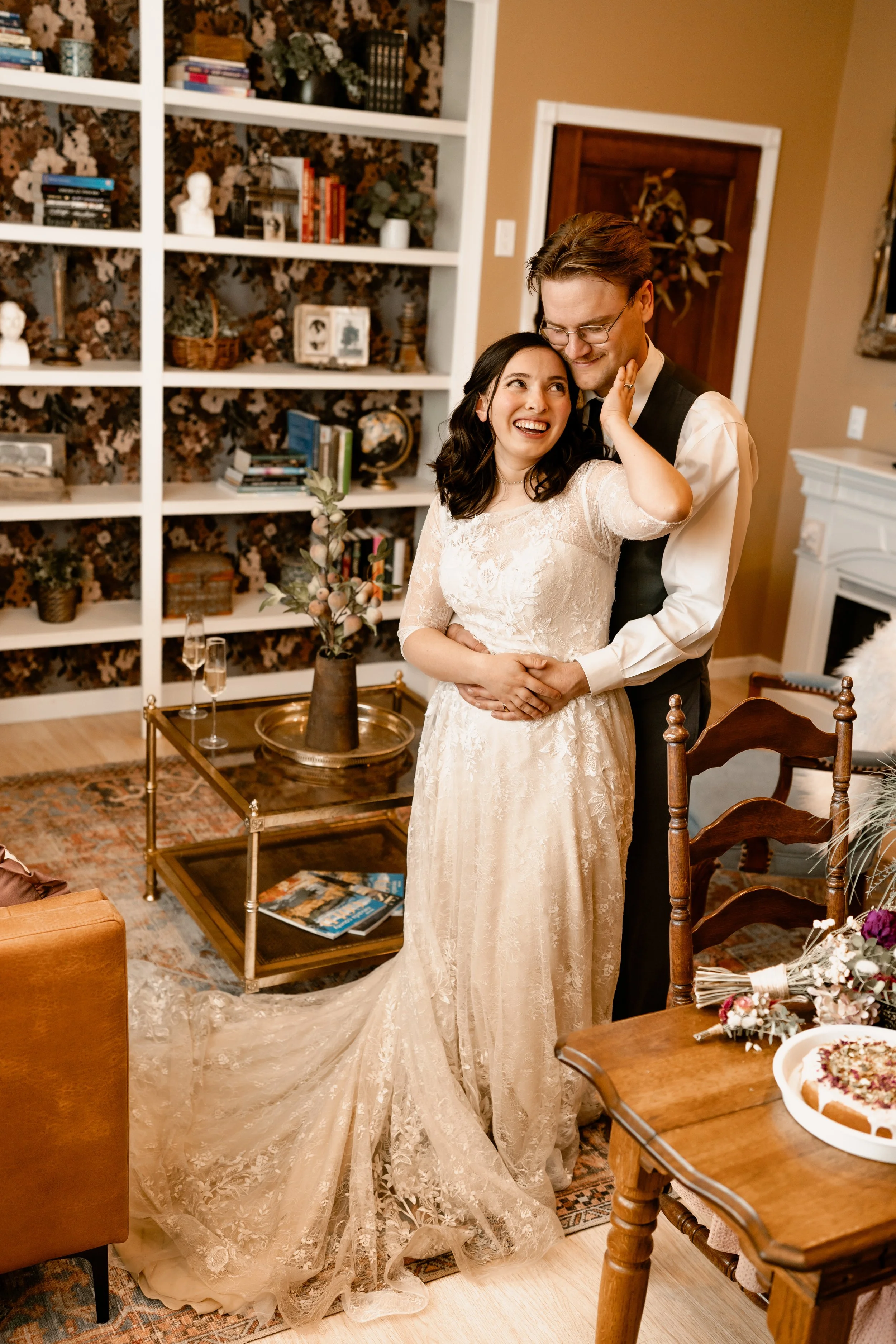 A bride and groom share a joyful moment in a cozy living room decorated with books, plants, and vintage decor. The bride wears a lace wedding gown, and the groom wears a vest and glasses. There is a table with champagne glasses, a sculpture bust, and