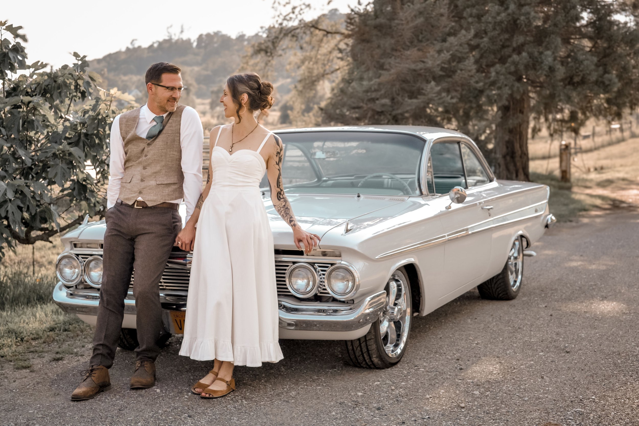 A couple standing in front of a vintage white car outdoors, holding hands and smiling at each other, with trees and hills in the background.