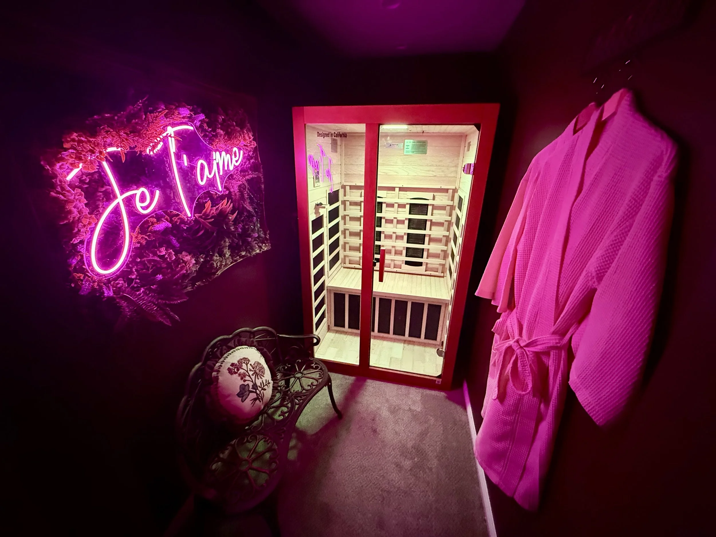Pink-lit room with a neon sign reading 'Je t'aime', a pink bathrobe hanging on the wall, a small decorative bench with floral cushion, and a sauna behind glass doors.