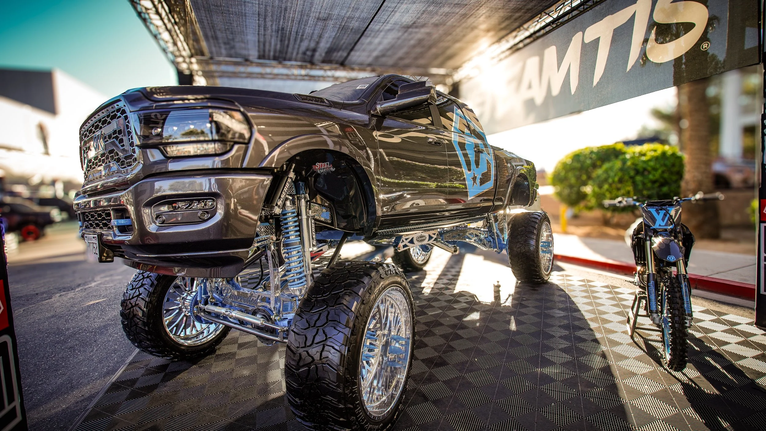 A replica monster truck and motorcycle with blue and black color schemes on display at an outdoor event, under a canopy with a sign reading 'AMT'.