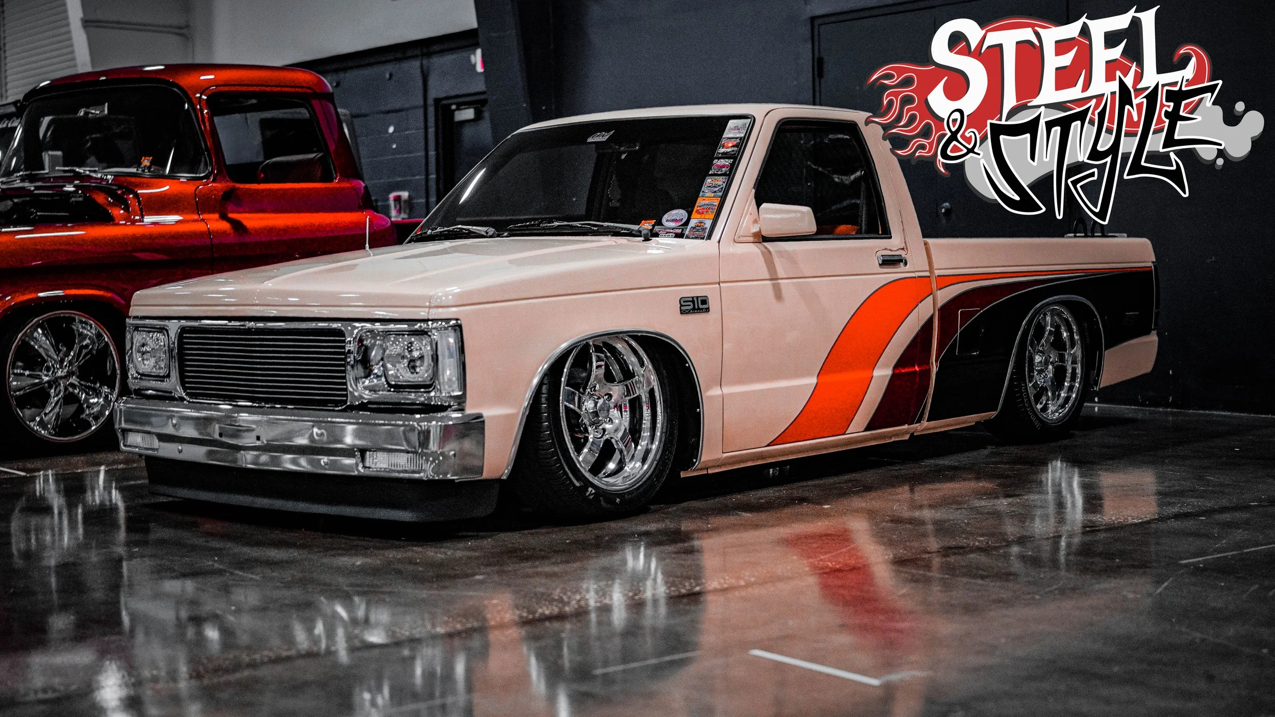 A custom vintage pickup truck with a tan, orange, and red paint job and chrome wheels, parked indoors next to another red vintage truck. The wall behind has a 'Steel & Style' graffiti logo.