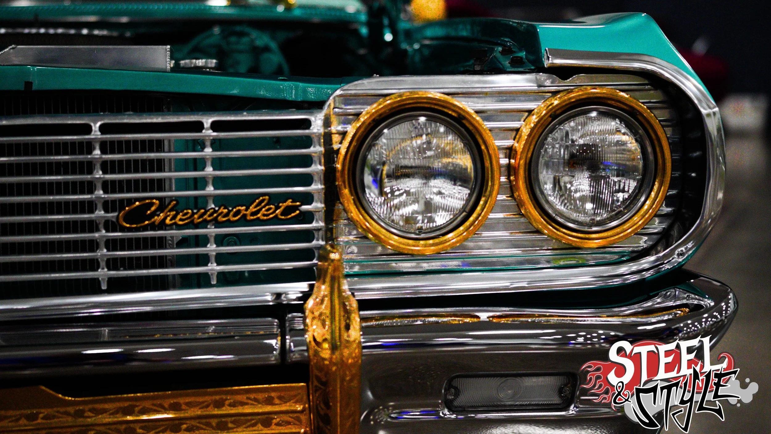 Close-up of the front of a vintage Chevrolet car with dual headlights. The car has a chrome grille with a Chevrolet badge and ornate golden accents, including golden rings around the headlights and a golden hood ornament. The image also features a lo