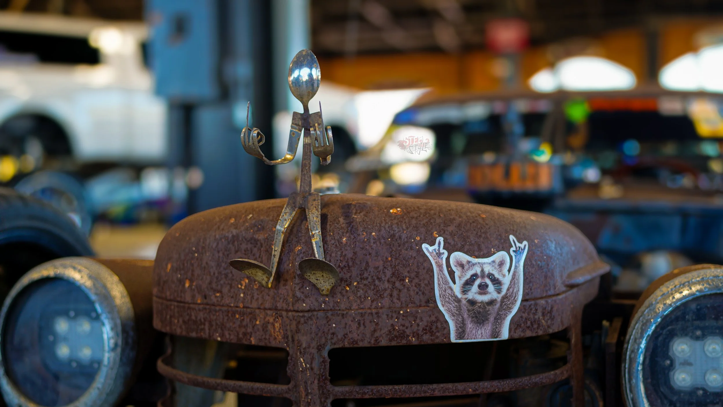 Rusty metal sculpture of a person made from utensils, sitting on a rusty surface, with a sticker of a raccoon raising both paws on the front.