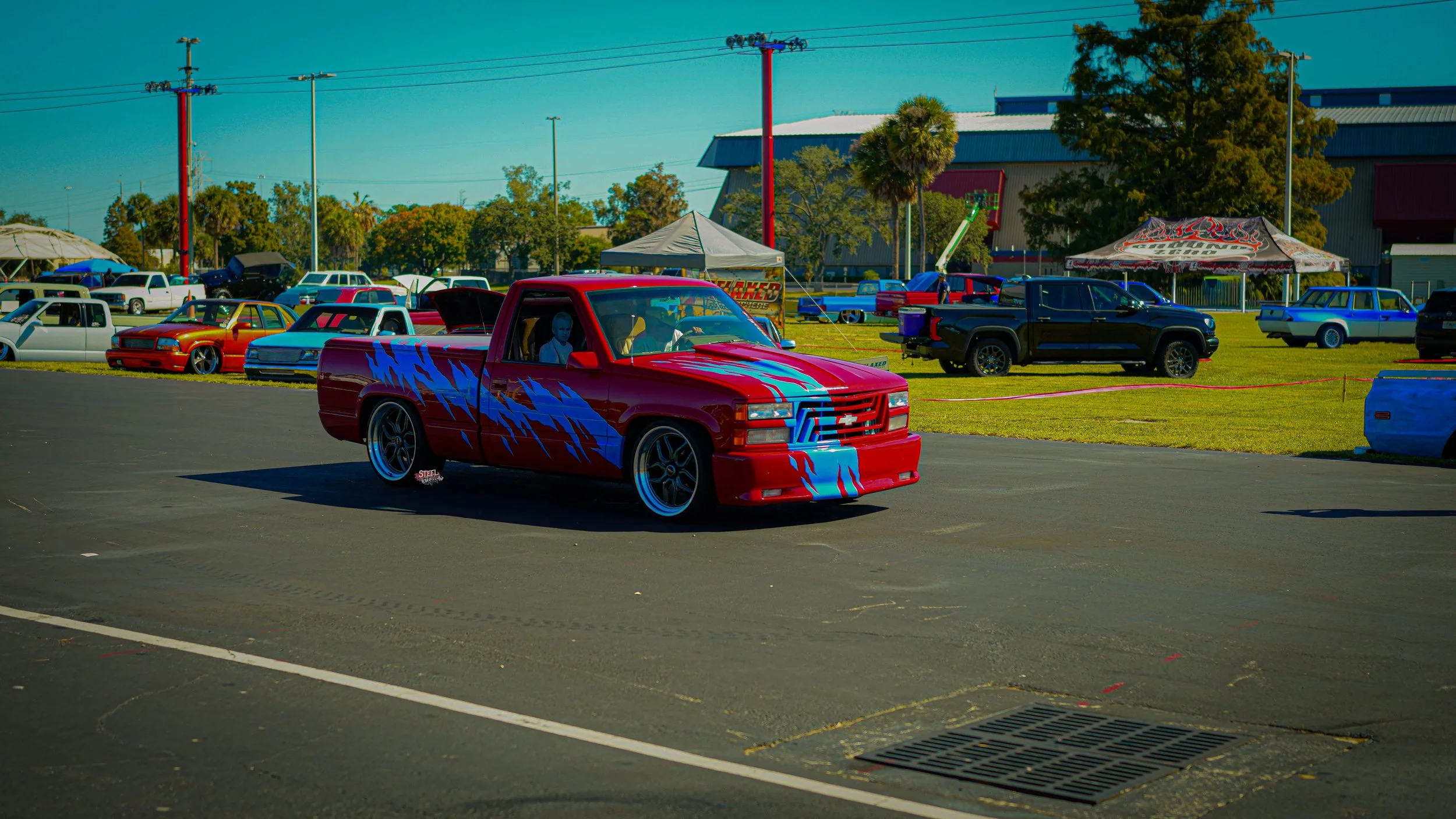 A red pickup truck with blue and purple lightning bolt graphics parked at a car show, with other vintage and custom cars parked behind it on a grassy field.