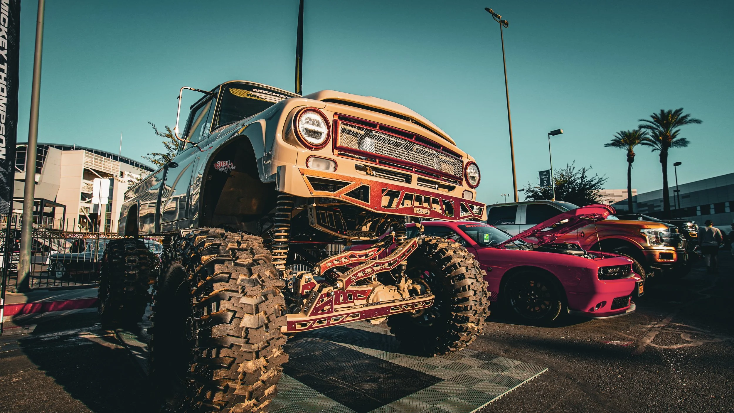 A heavily modified monster truck with very large tires and custom suspension is parked at an outdoor car show, alongside a red sports car with its hood open and a black SUV, with palm trees and buildings in the background during daylight.