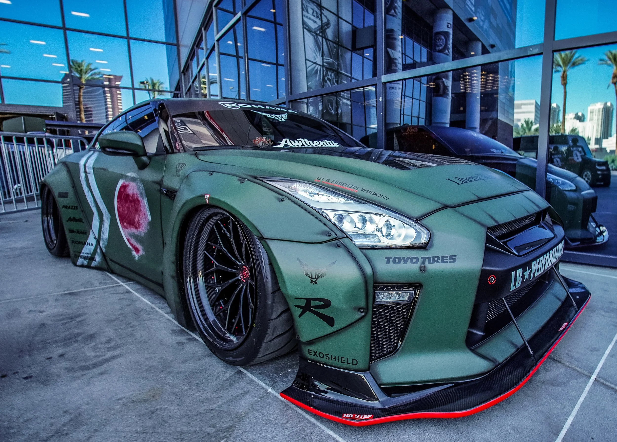 A sleek, customized racing car with a matte green finish parked outside a modern glass building. The car features various sponsor decals, black racing wheels, and a prominent front splitter with red trim.