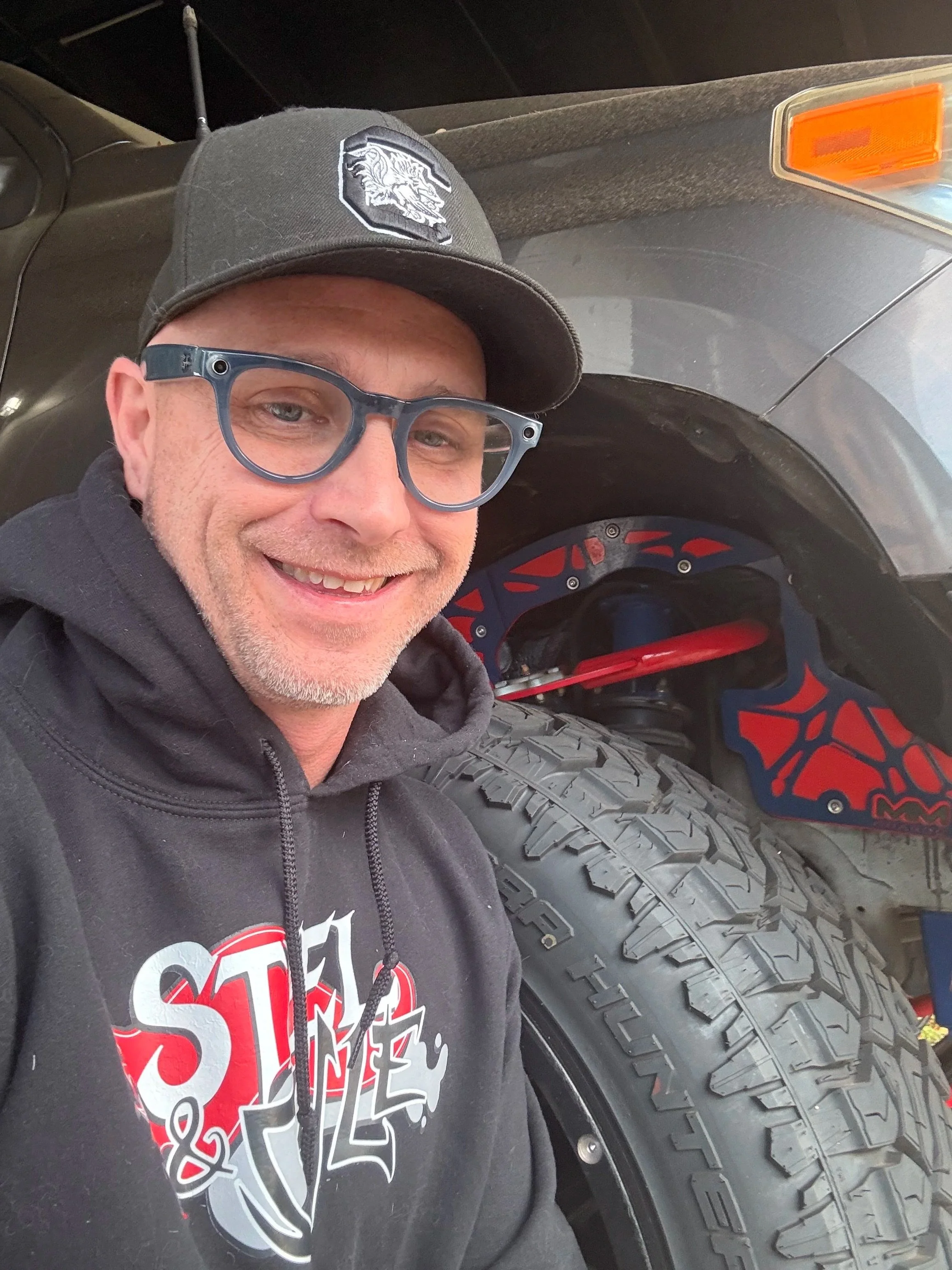 A man smiling, wearing sunglasses, a black cap with a lion logo, a black hoodie, and a black T-shirt with red and white graphics. He is sitting next to a rugged off-road vehicle with a large tire featuring deep treads, a black fender, and a wheel with red and black accents.