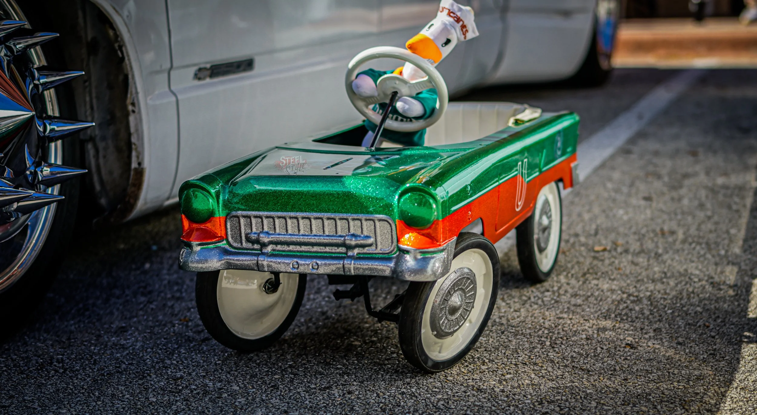Toy ride-on car modeled after a vintage green and orange car with a white steering wheel, parked on asphalt next to a silver vehicle.