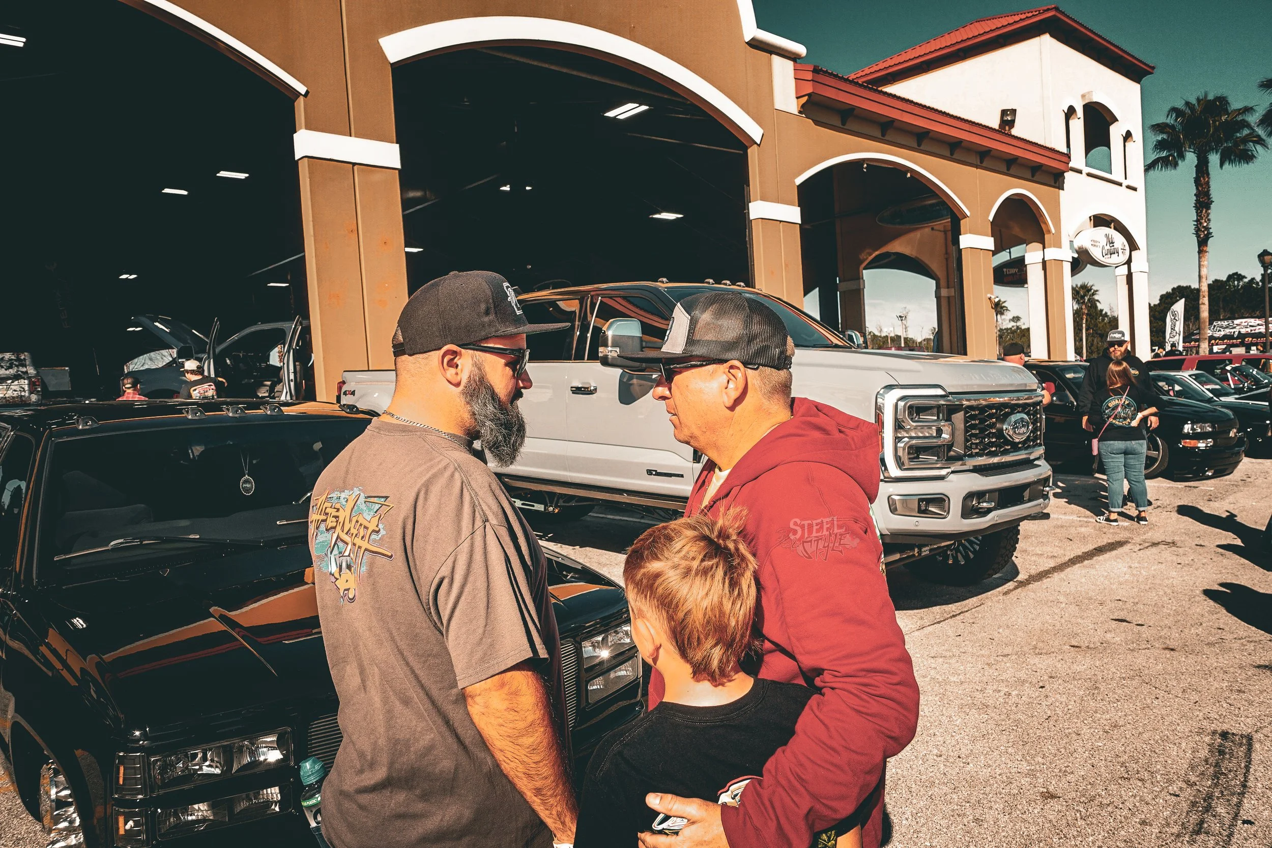 Three people standing close together outdoors in front of a building with parked cars and palm trees. Two men and a child are engaging in conversation, with one man holding the child's arm.