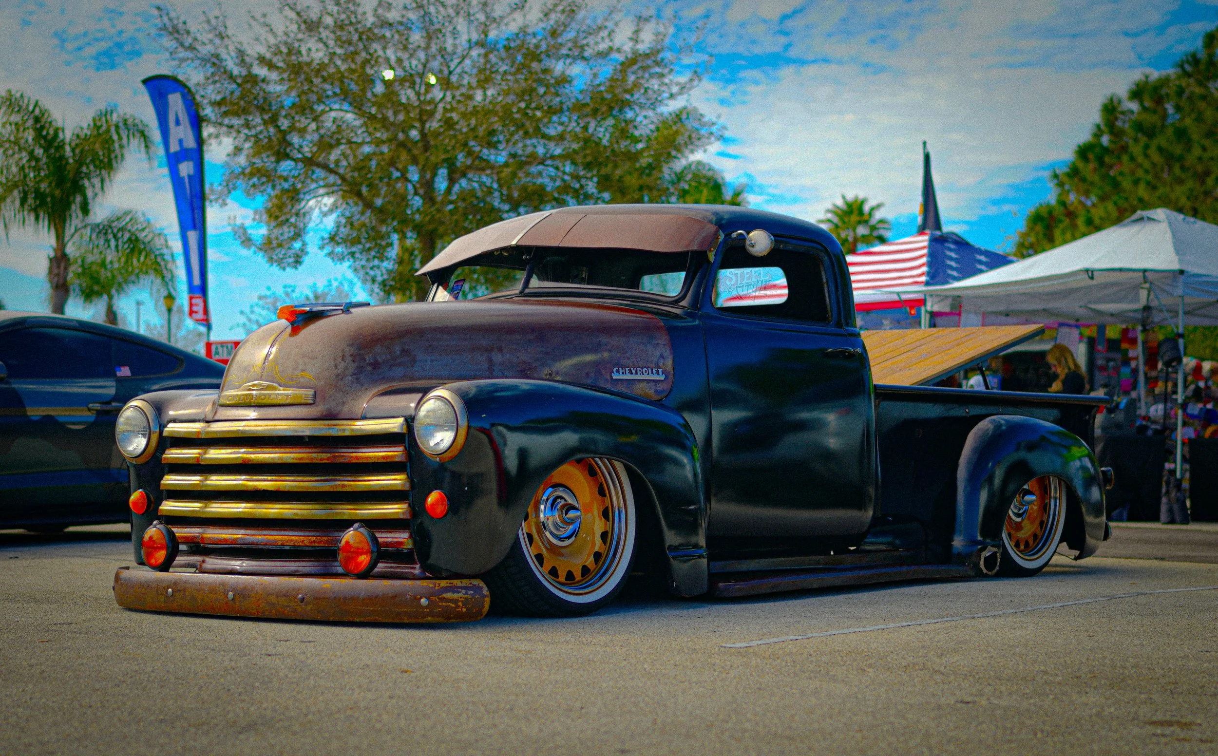 A vintage black pickup truck with a rusted hood and lowered stance parked at a car show, with tents and trees in the background.