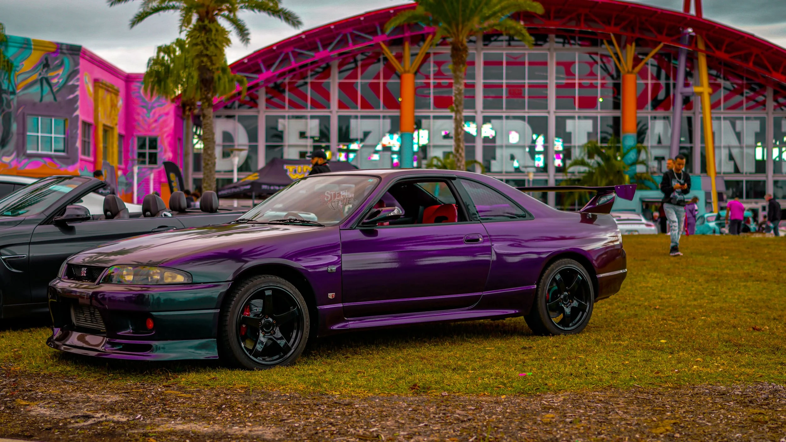 A purple sports car parked on grass at a car show in front of a colorful building with palm trees.
