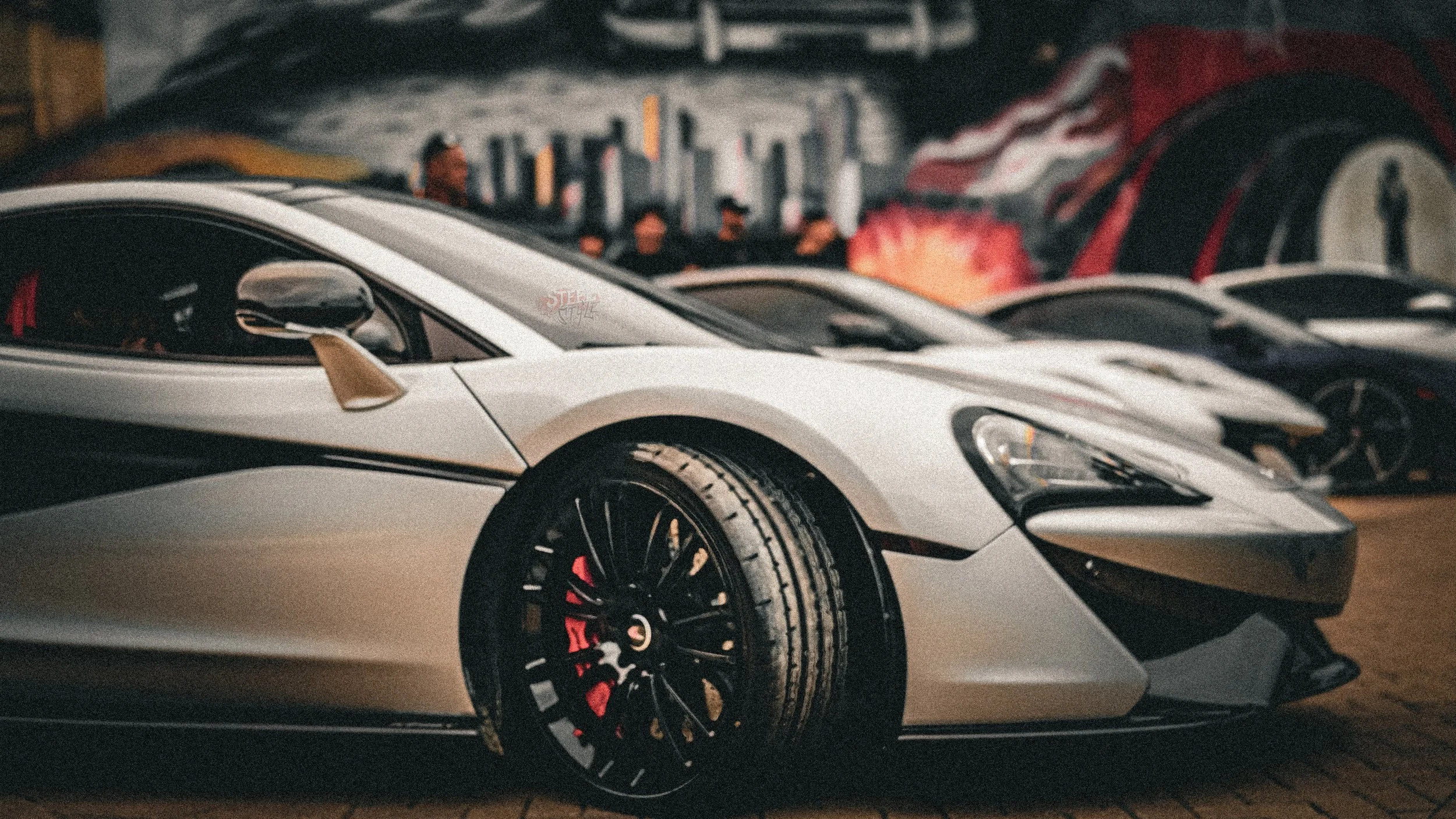 A line of luxury sports cars parked indoors, featuring a white McLaren with black wheels and red brake calipers in the foreground.