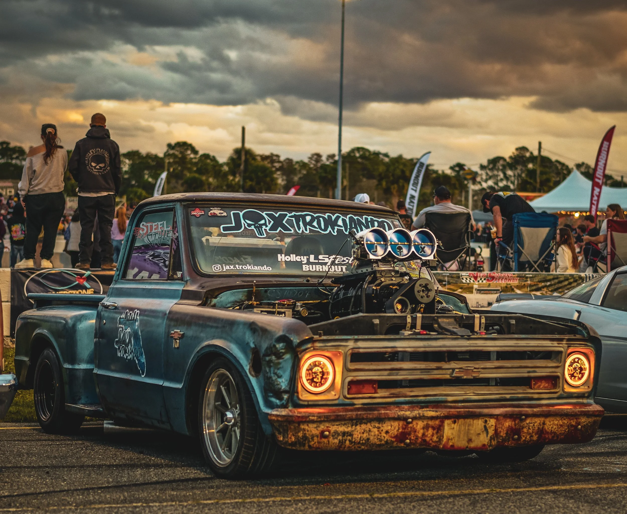 A vintage Chevrolet pickup truck with a rusted, weathered body and exposed engine, parked at an outdoor car show during sunset with a crowd of people and tents in the background.