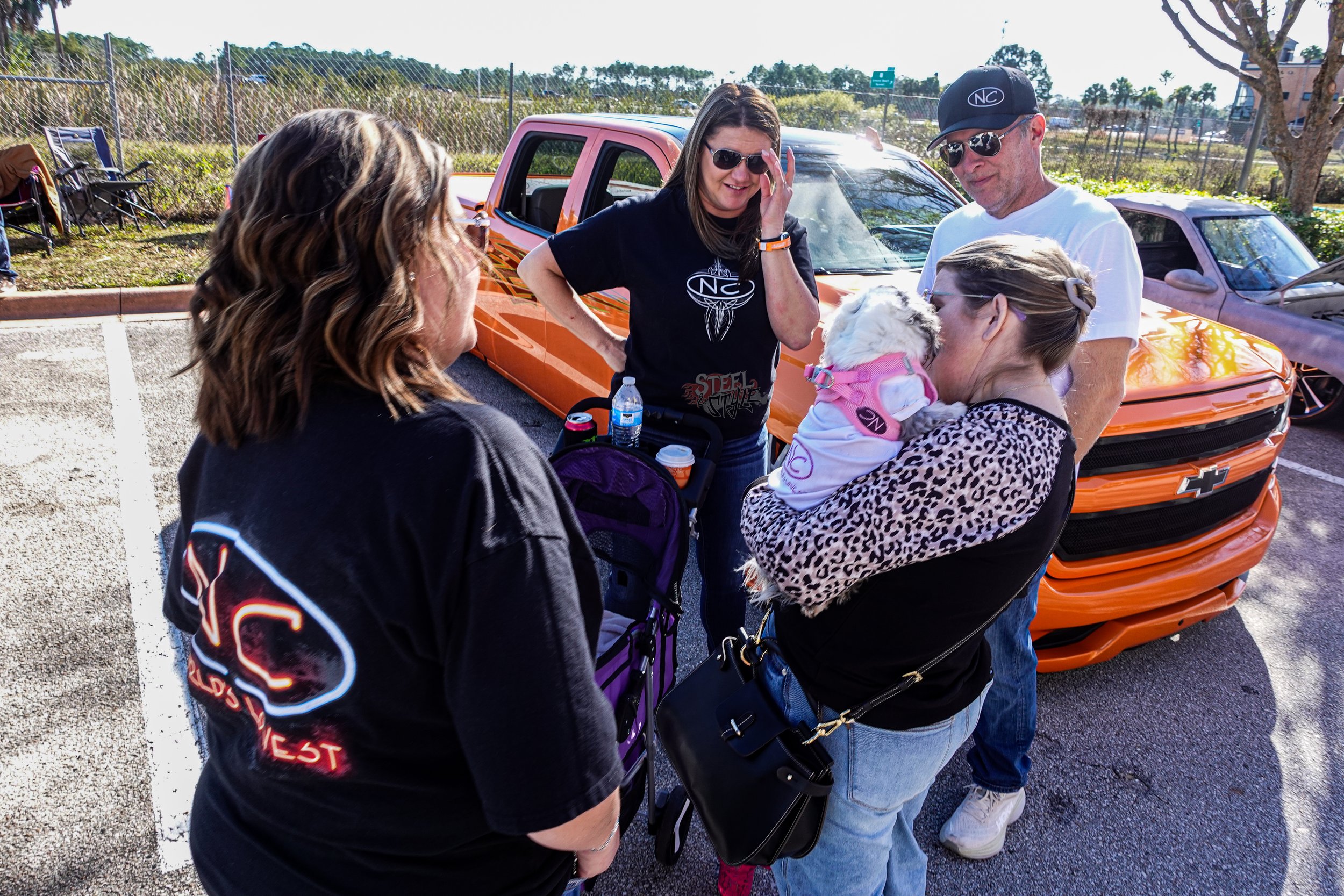 Four people standing near a bright orange Chevrolet Camaro, one woman holding a small dog in her arms, in a parking lot with a chain-link fence and trees in the background.