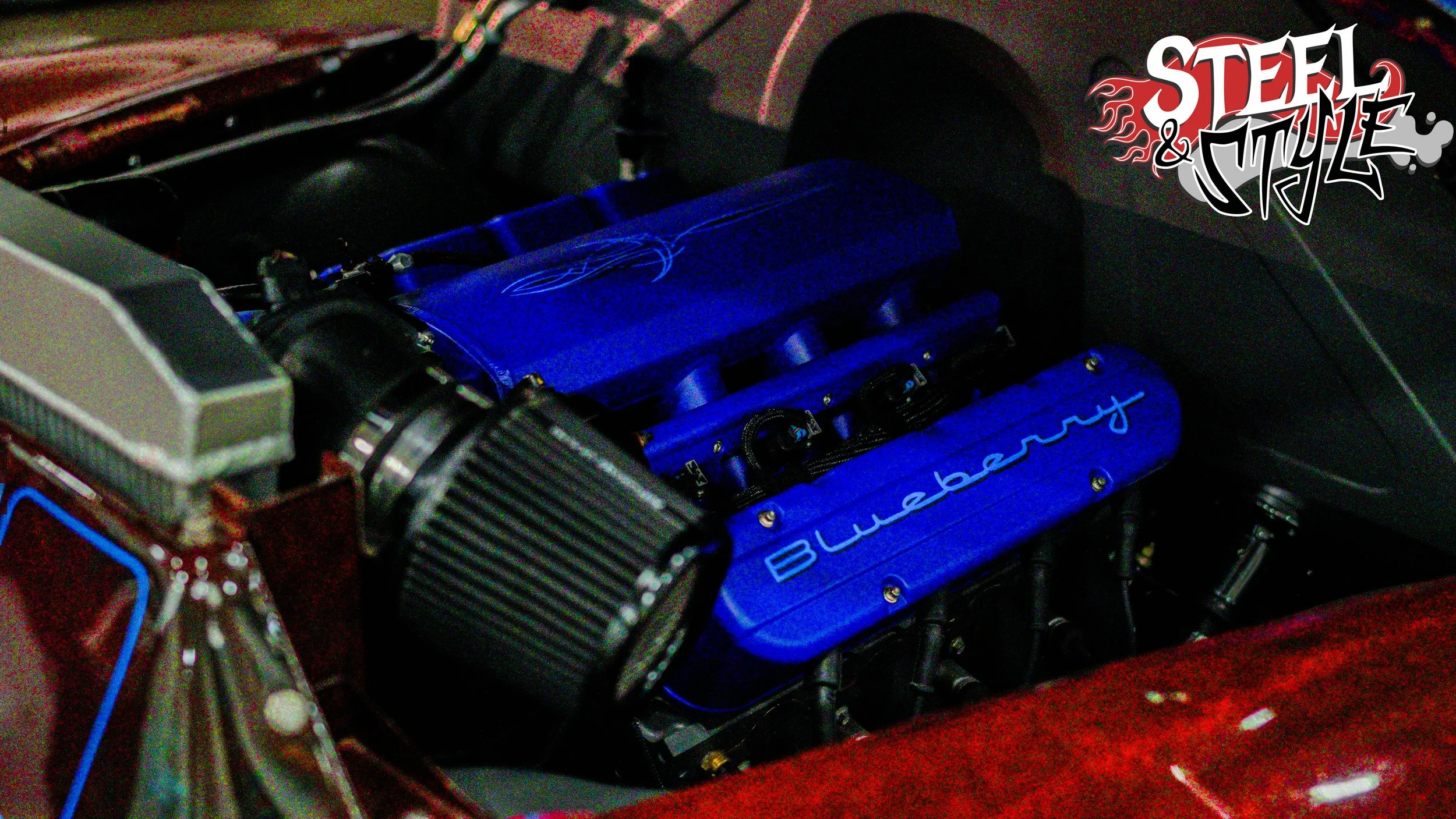 Close-up view of a car engine with blue valve covers labeled 'Blueberry' and 'Steel & Style' logo in the top right corner