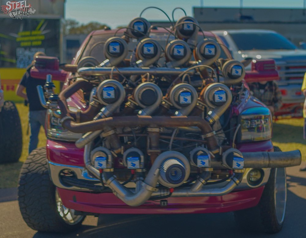 Front view of a modified red car with a large, exposed supercharger and multiple attachment points on the engine, likely at a car show or racing event.