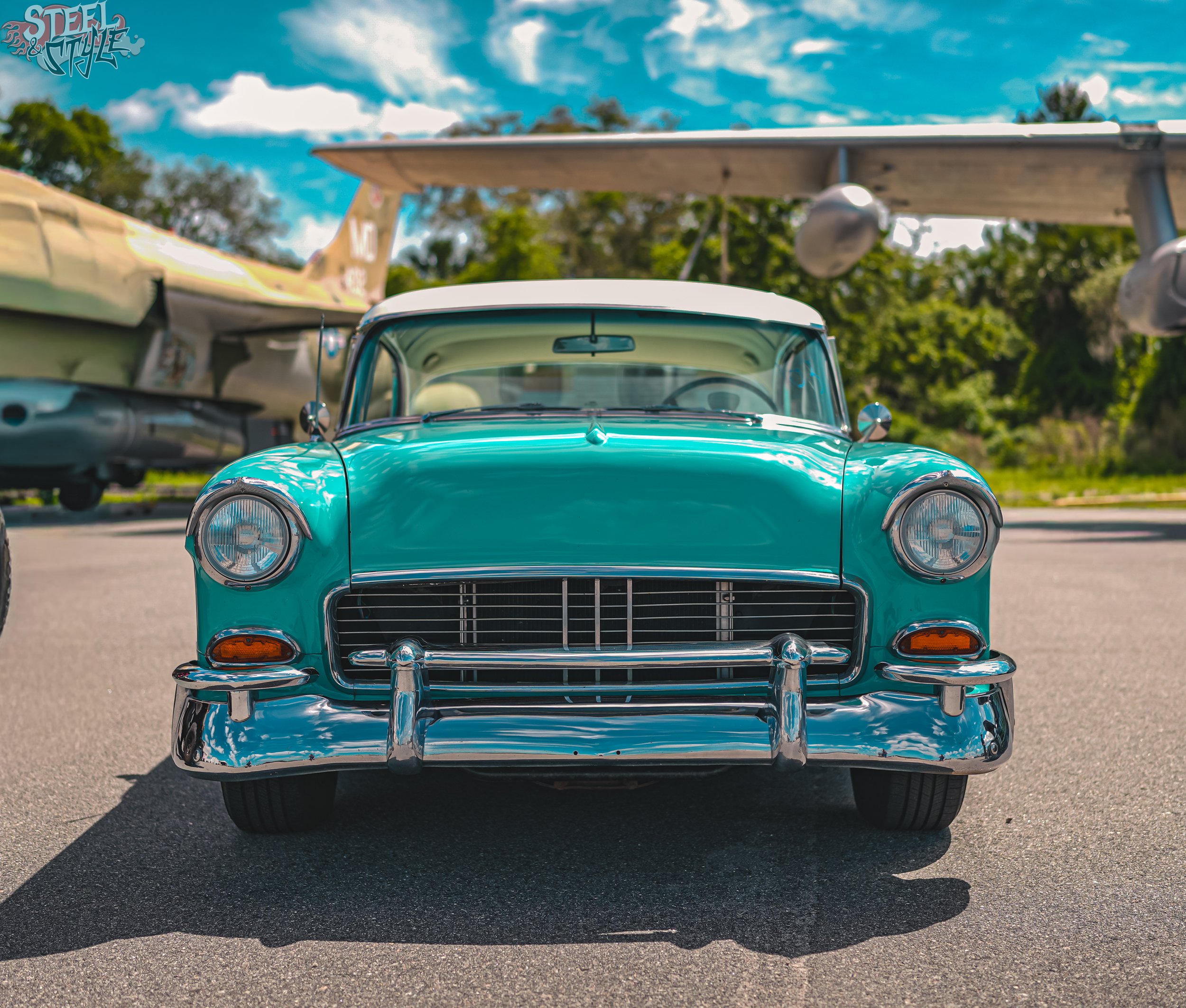 Front view of a vintage turquoise car parked outdoors with aircraft in the background under a partly cloudy sky.