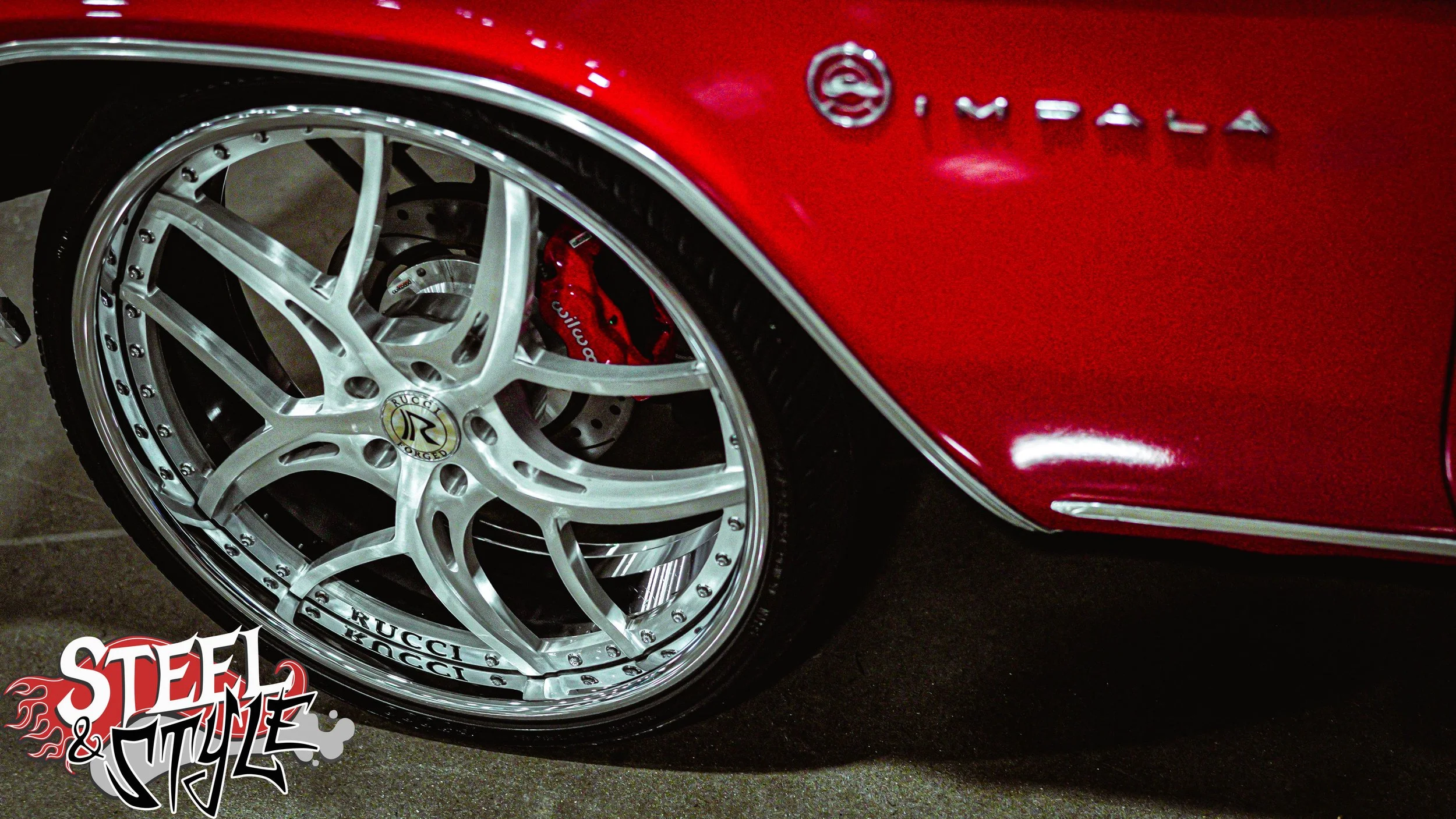 Close-up of a red sports car's wheel and brake caliper. The wheel has a silver, multi-spoke design with the RUCKI logo, and the brake caliper is painted red with the brand name written on it. The car has a shiny red body with chrome accents and the w
