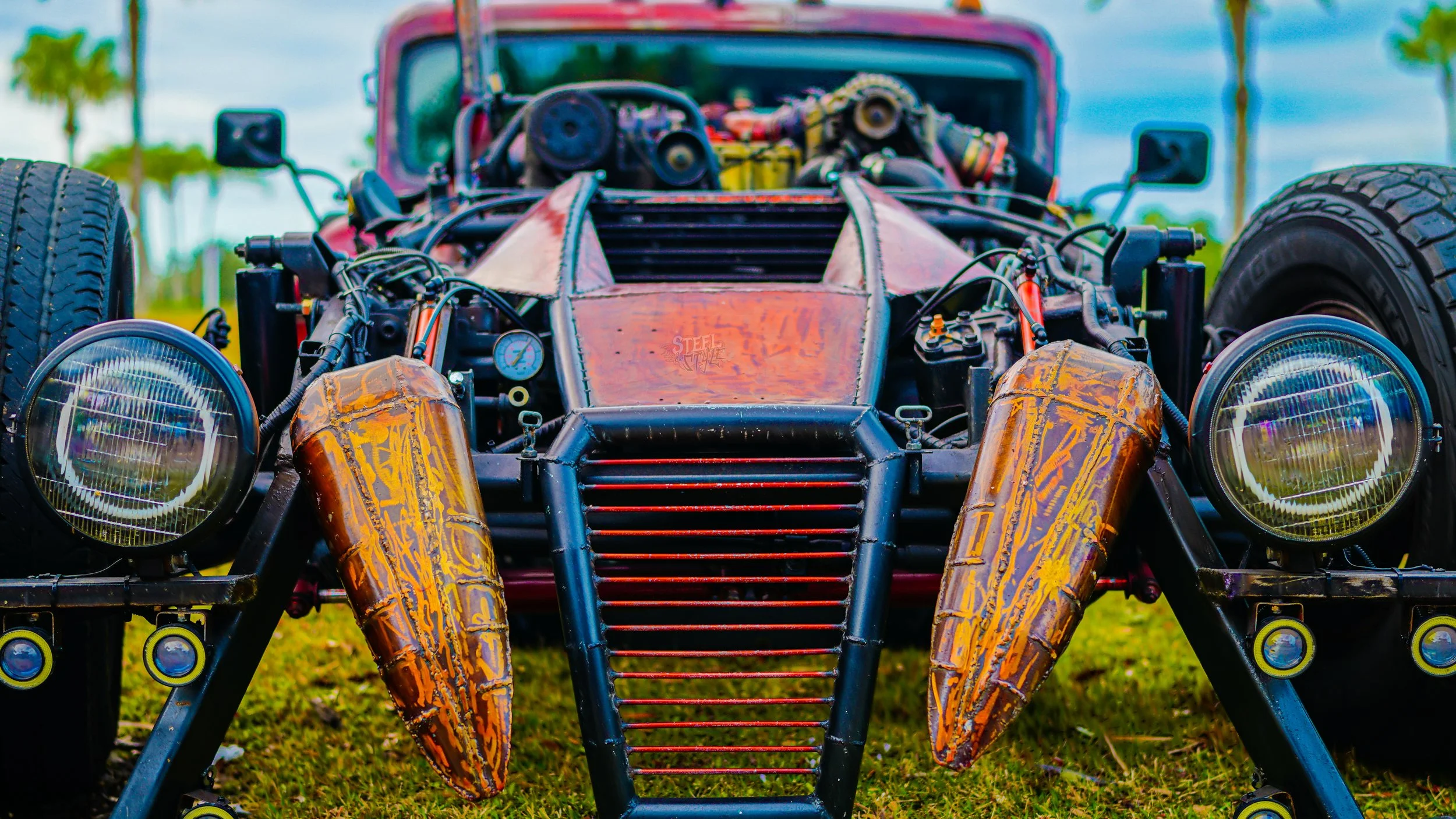 Close-up of a vintage race car with exposed engine parts, large round headlights, rusty body panels, and a black grille, parked on grass.