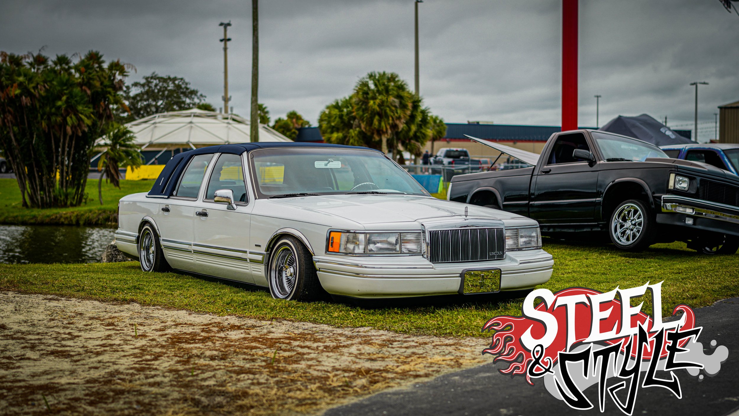 A white vintage Lincoln car with a dark blue roof parked on grass near a pond at a car show, with other classic vehicles and trees in the background, and a "Steel & Style" logo in the lower right corner.
