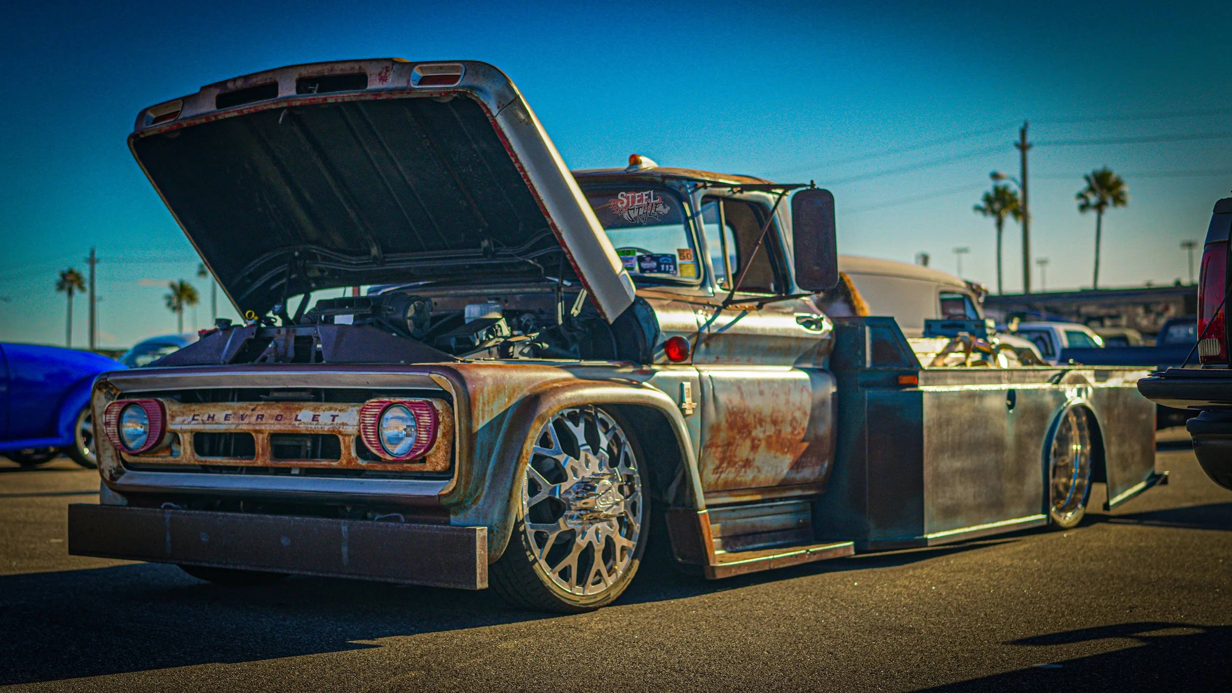 A vintage Chevrolet race truck with a rusty, weathered look, wide racing tires, and open hood showing the engine, parked on a lot with palm trees and other cars in the background during sunset.