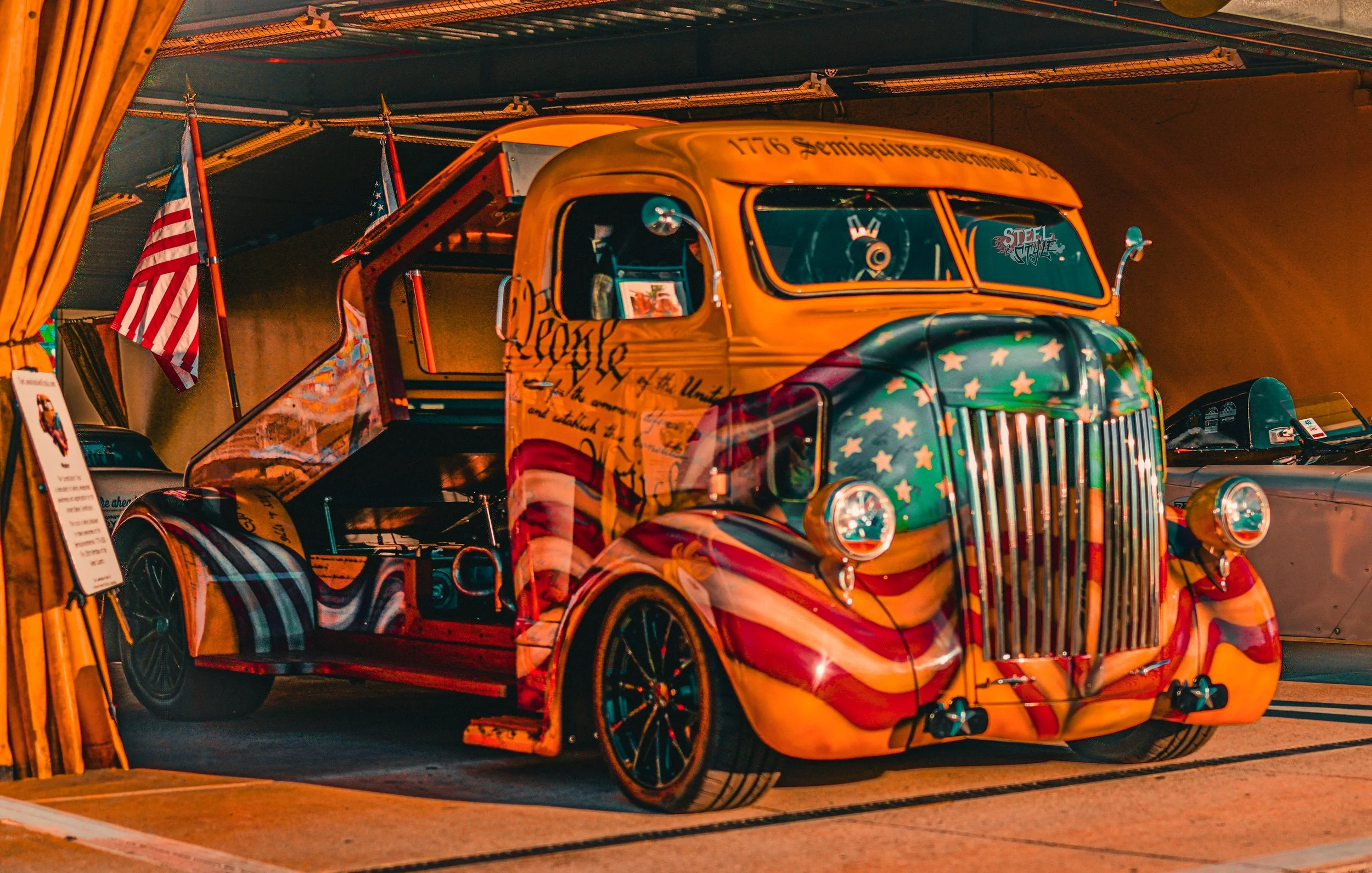 A vintage truck painted with an American flag design and stars on the hood, displayed indoors at an exhibition with American flags in the background.