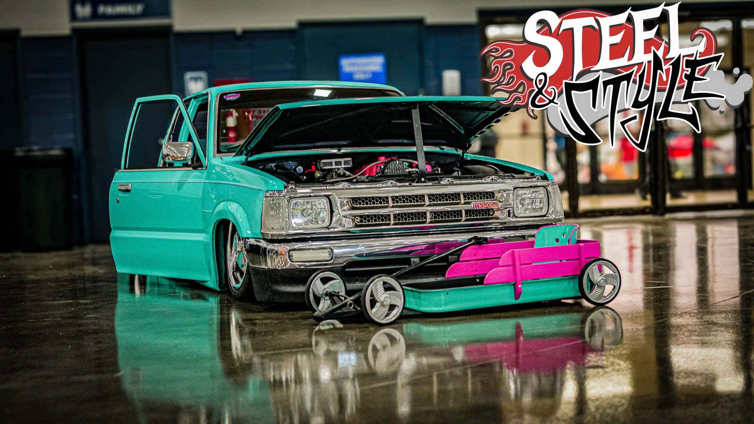 A small turquoise toy car with a pink bumper, with its hood open revealing the engine. The toy car is equipped with a skateboard in front of it, and there's a colorful graffiti-style text overlay reading "Steel & Style" in the upper right corner.