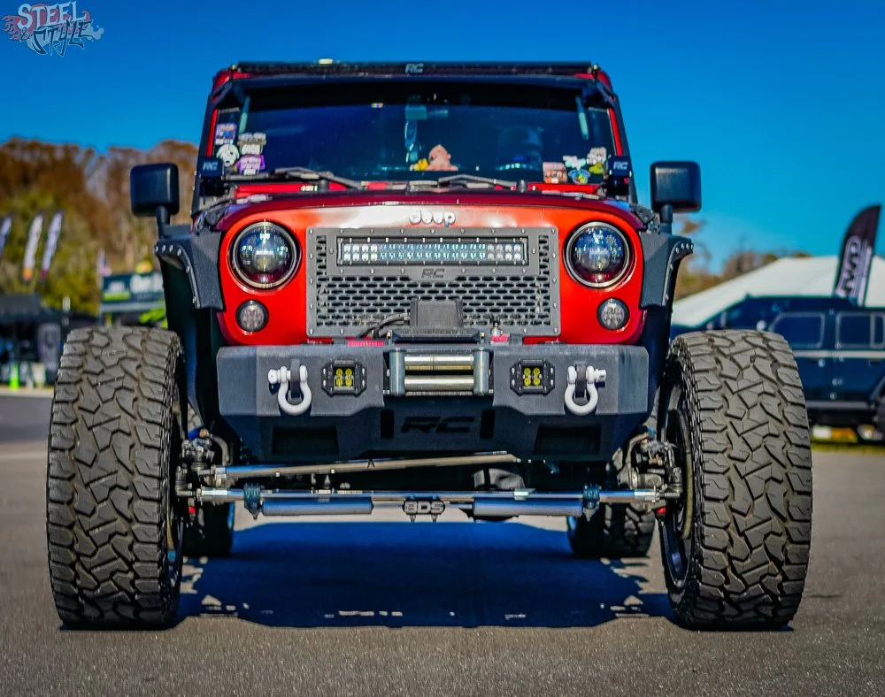 Front view of a red off-road Jeep with large tires, aftermarket bumper, and LED light bar, parked outdoors at a motorsport event.
