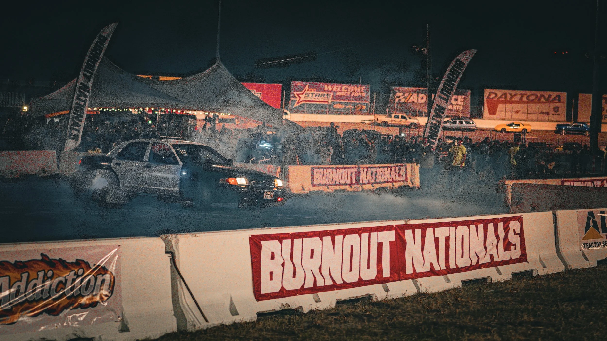 Night scene at a drag racing event at Burnout Nationals, featuring a silver racing car on the track with smoke and tire burnout. The background includes a crowd of spectators, tents, banners, and billboards.