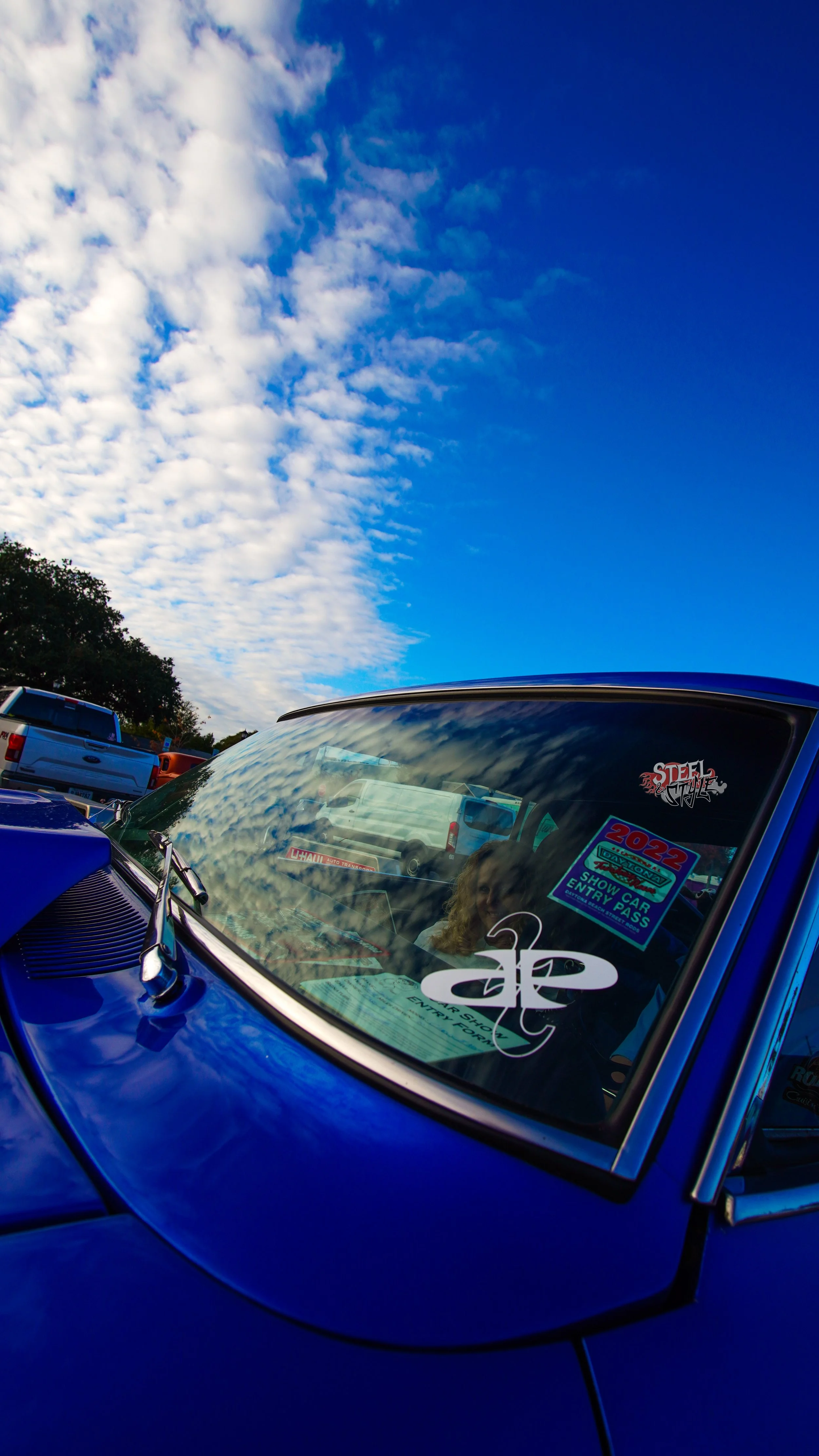 Close-up of a blue vintage car windshield with reflections of clouds, showing stickers including a 2022 show car entry pass, a Tattoo logo, and a person inside.