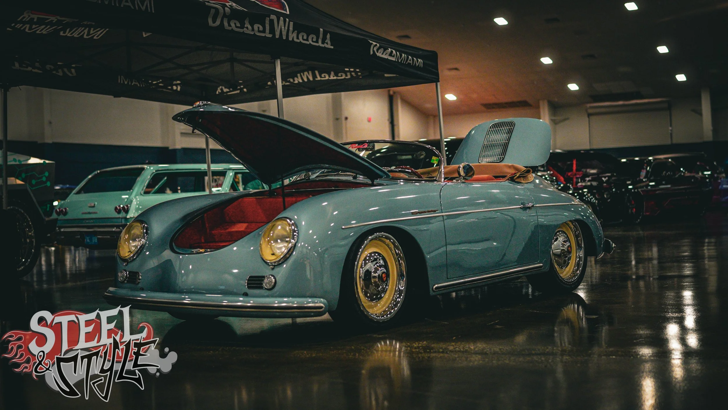 A light blue vintage convertible car with yellow hubcaps on display at an indoor car show. The car has its hood open, revealing the engine, and the interior is visible, showing red upholstery. Other classic cars are in the background under a tent wit