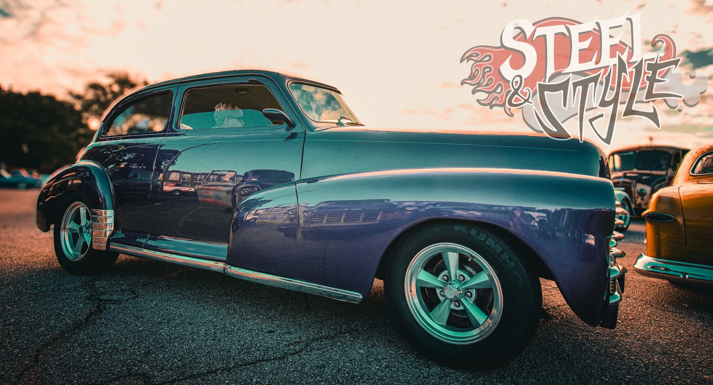 Vintage blue classic car parked in a lot at sunset with other cars visible in the background, and a stylized 'Steel & Style' logo in the top right corner.