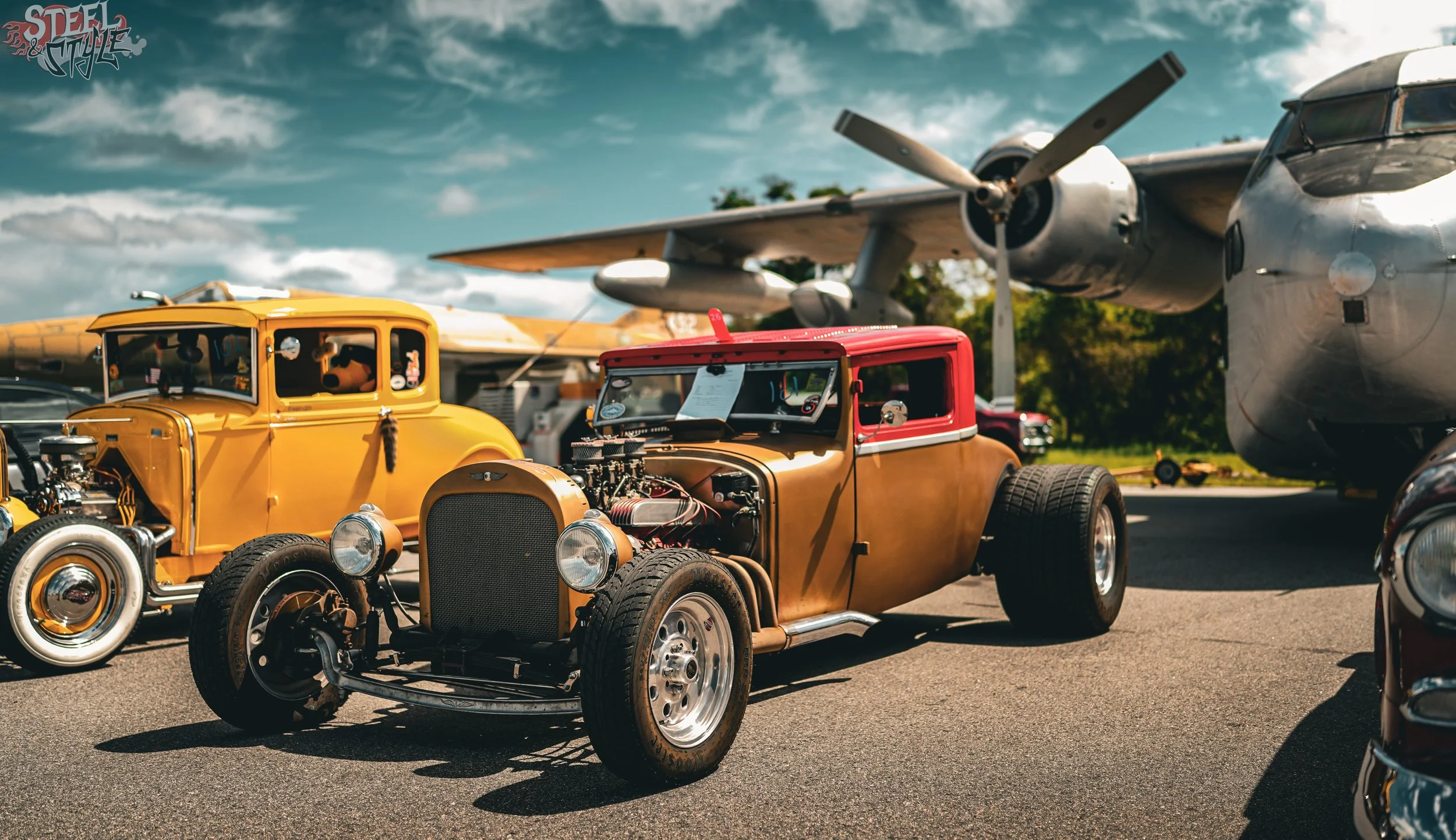 A collection of vintage cars displayed outdoors, with some cars showing exposed engines, and an airplane in the background under a partly cloudy sky.