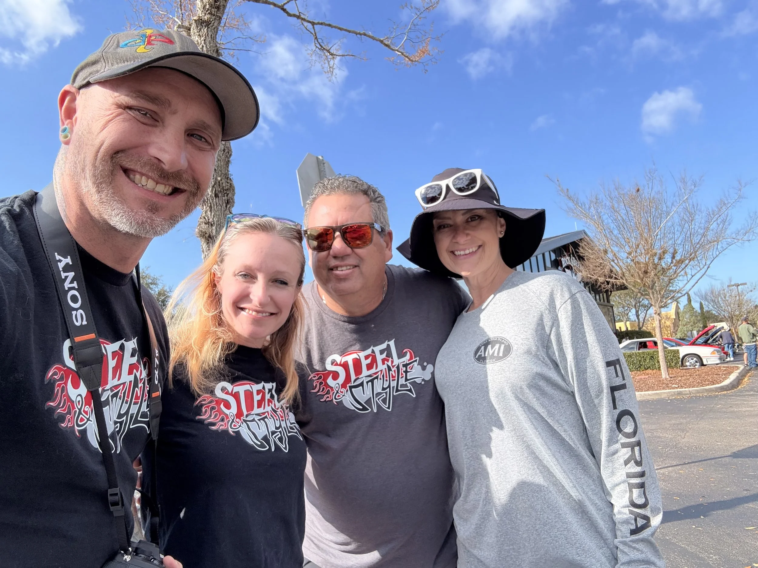 A group of five smiling people posing for a photo outdoors against a clear blue sky, with trees and cars in the background. Four of them are wearing matching black shirts with Red and Grey text, and one person is wearing a grey hoodie with black sleeves. Two individuals are wearing sunglasses, and one is wearing a hat with sunglasses on top.