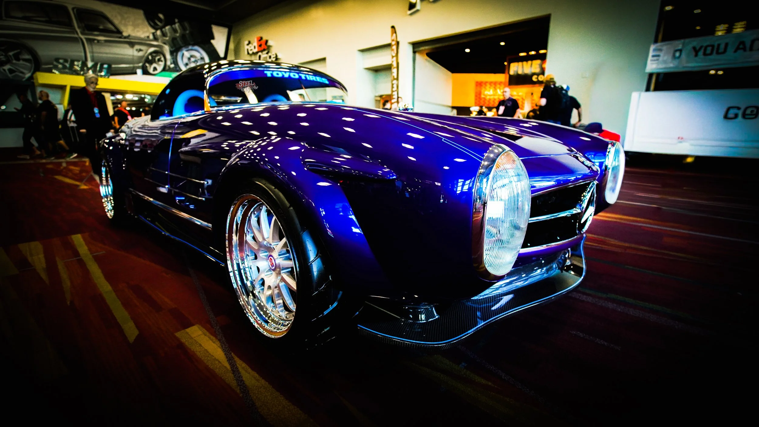 A shiny blue vintage sports car with chrome wheels displayed indoors at an auto show, surrounded by people and other exhibits.