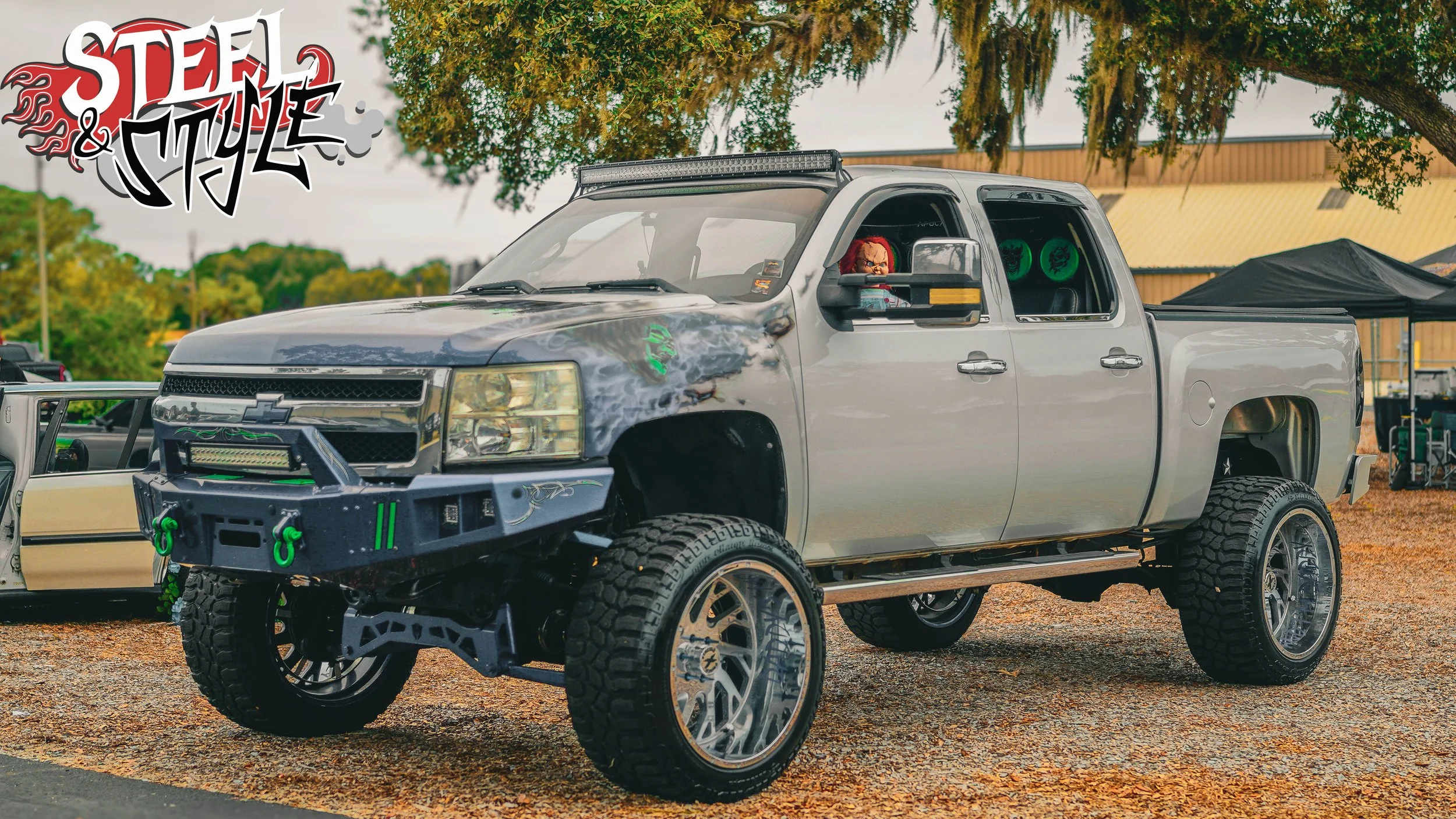 Modified gray Chevrolet pickup truck with custom wheels, off-road tires, and green accents parked outdoors under a tree at a car event. The truck has graffiti-style graphics, a light bar on the roof, and a small figurine of a child in the driver's se