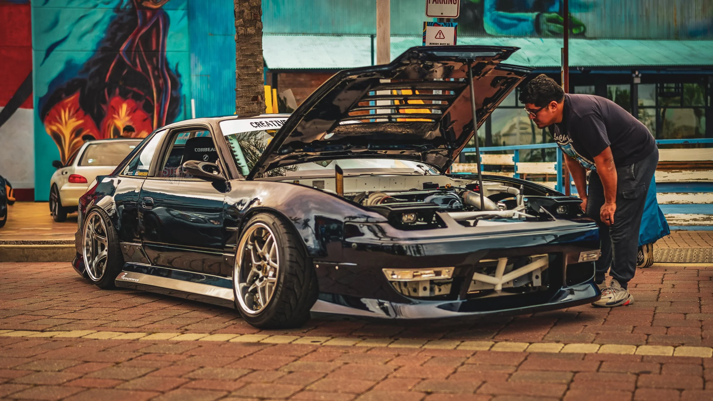 A man is inspecting a black sports car with its hood open, parked on a brick-paved area. There are two other cars in the background and colorful murals and signs on a building wall.