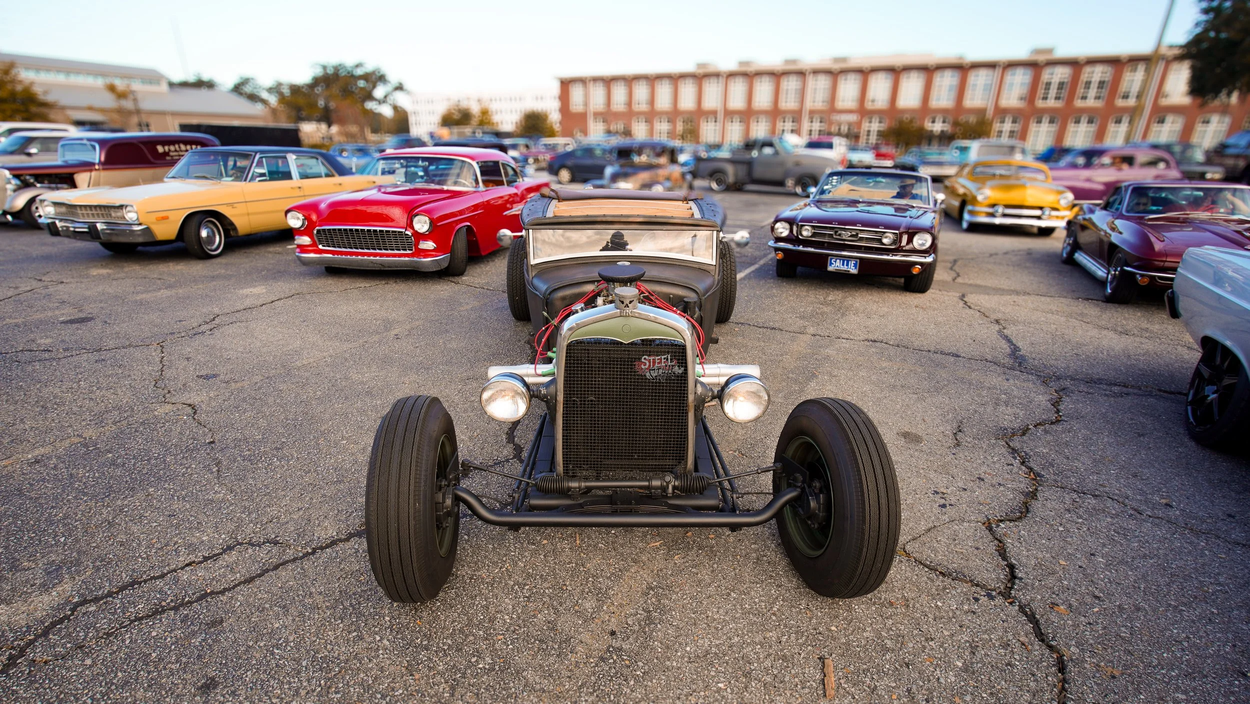 A collection of vintage cars parked on a cracked asphalt lot, with a clear sky and a building in the background.